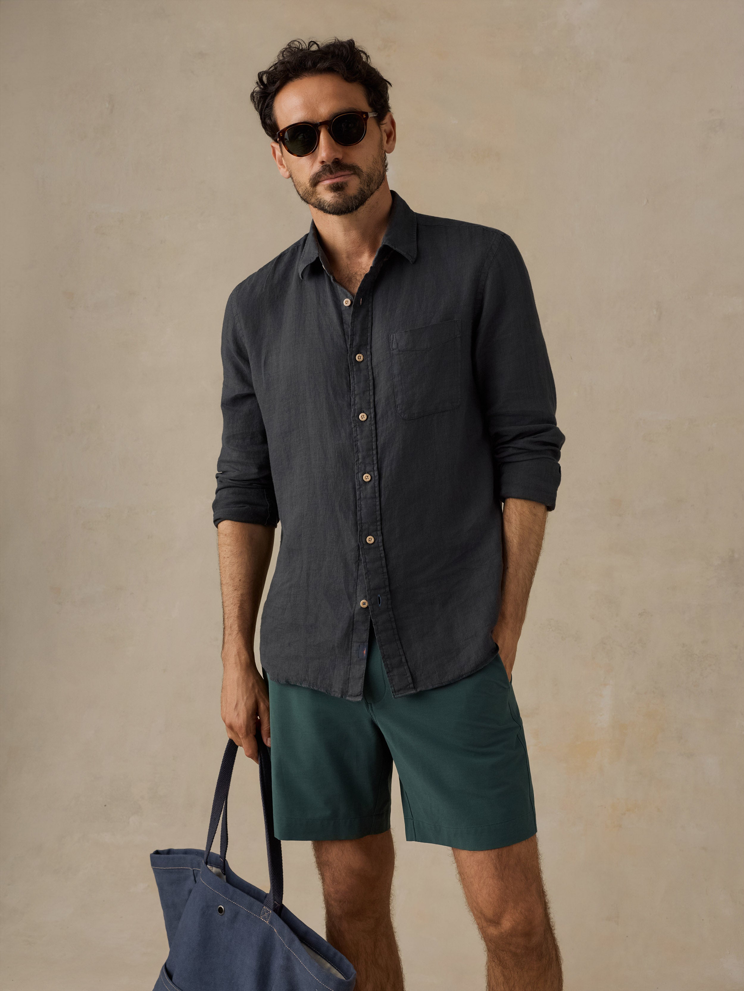 A man wearing a dark charcoal gray linen button-down shirt, teal green shorts, and sunglasses, holding a navy blue tote bag against a textured beige background.