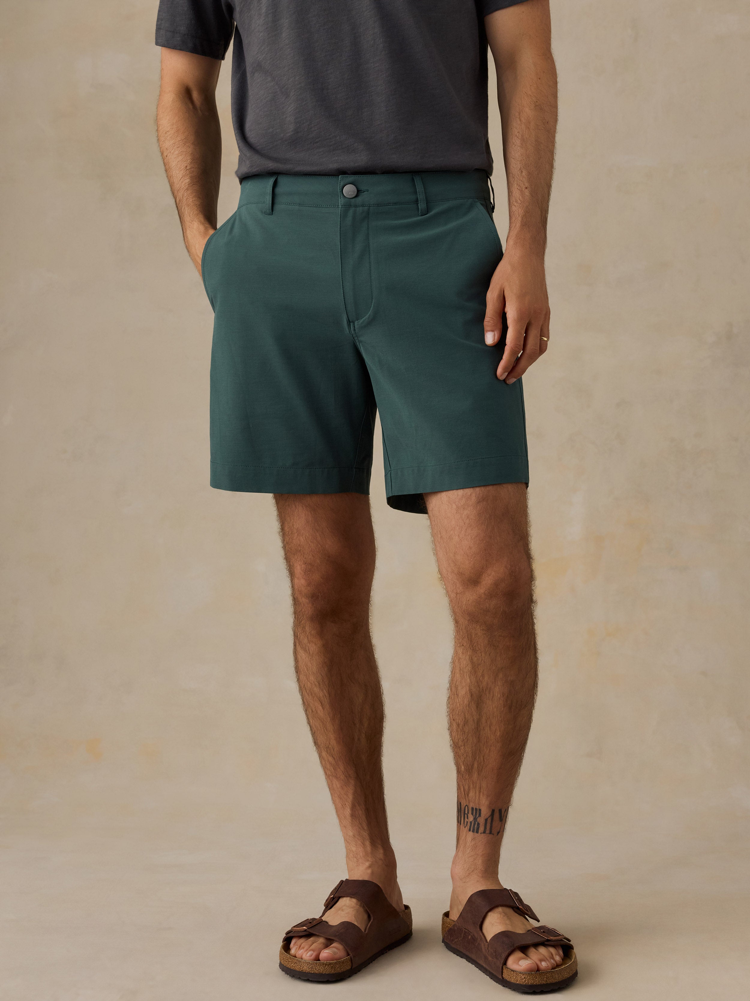 A man wearing dark teal-green chino shorts, a dark gray t-shirt, and brown slide sandals, standing against a textured beige studio backdrop.