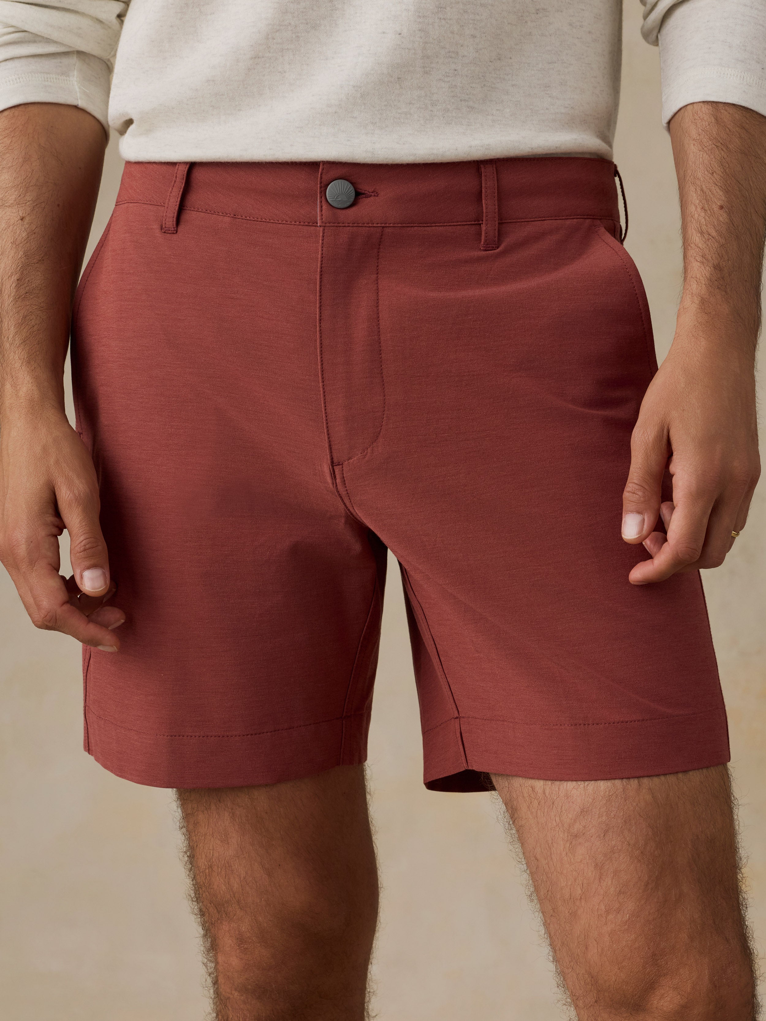A close-up of a man wearing rust-colored chino shorts with a cream knit top, shown from the waist to mid-thigh against a warm beige background.
