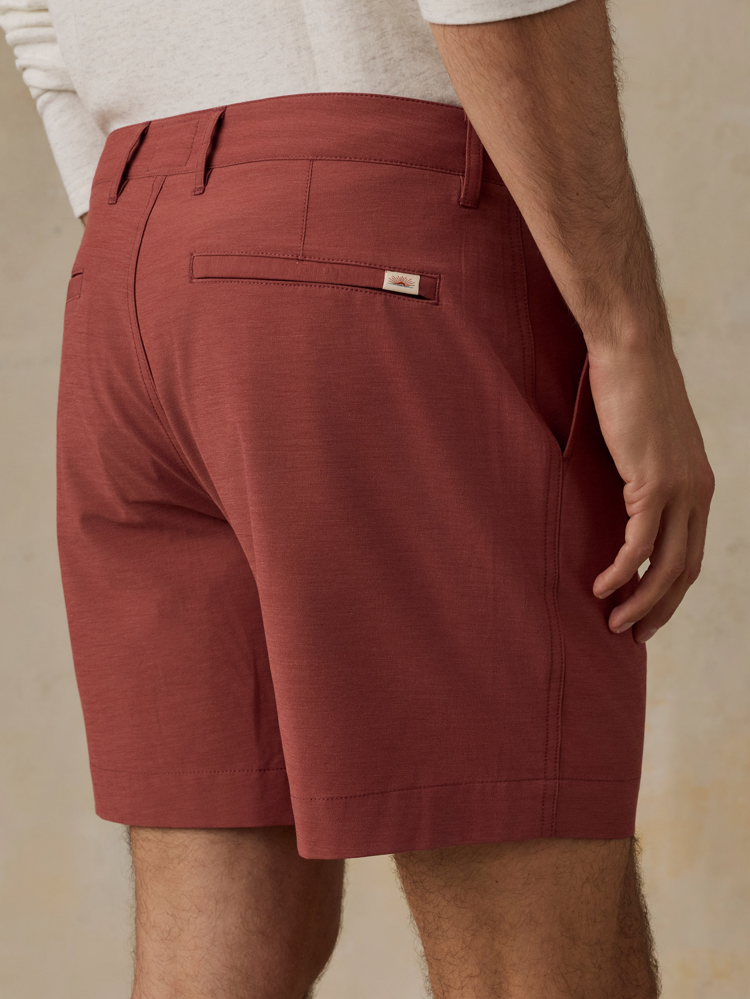 A close-up back view of a man wearing rust-colored chino shorts with a white shirt partially visible at the top, set against a neutral beige background.