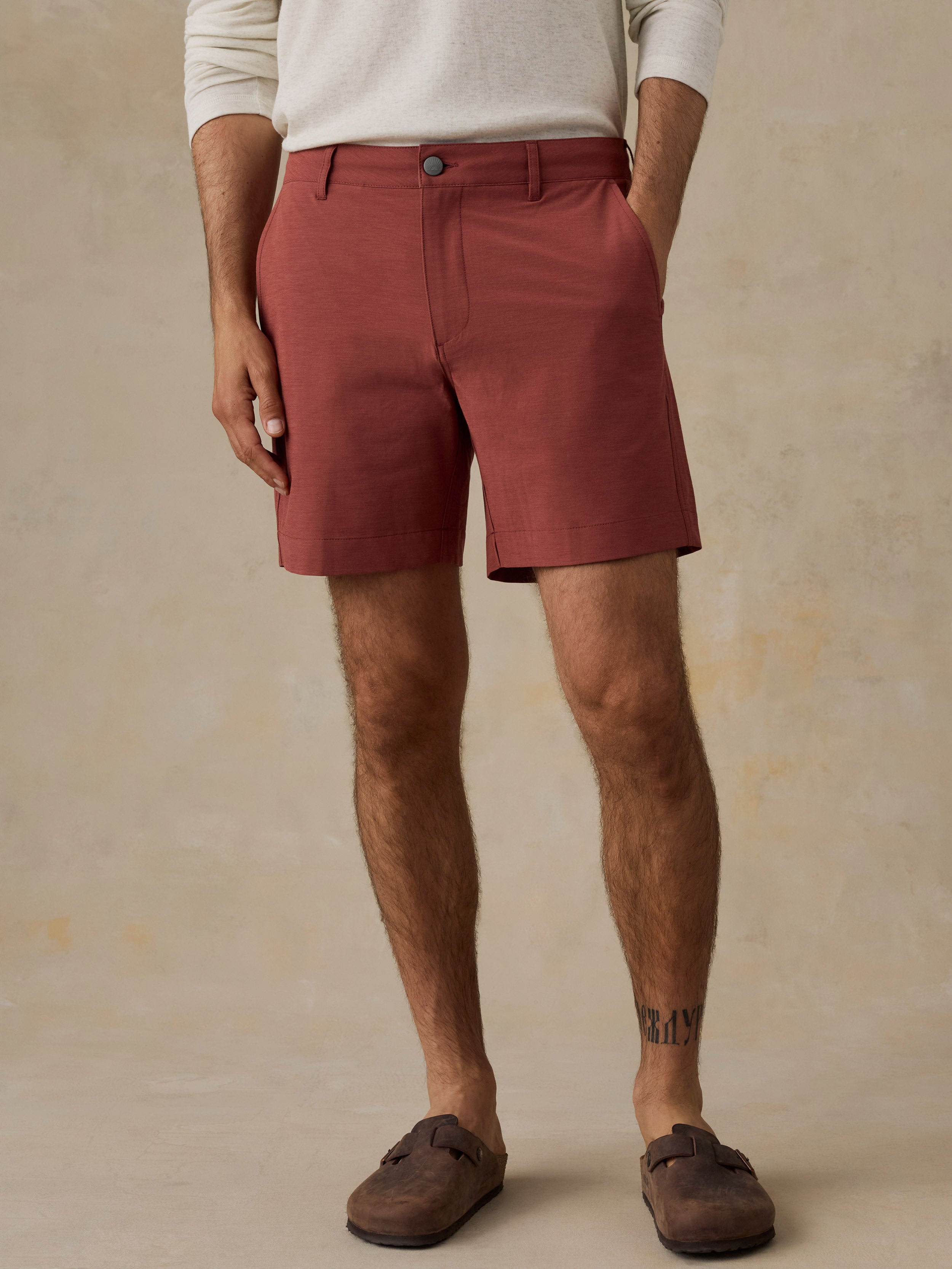 A man wearing rust-colored chino shorts, a cream long-sleeve shirt, and brown suede mules, standing against a textured beige studio backdrop.