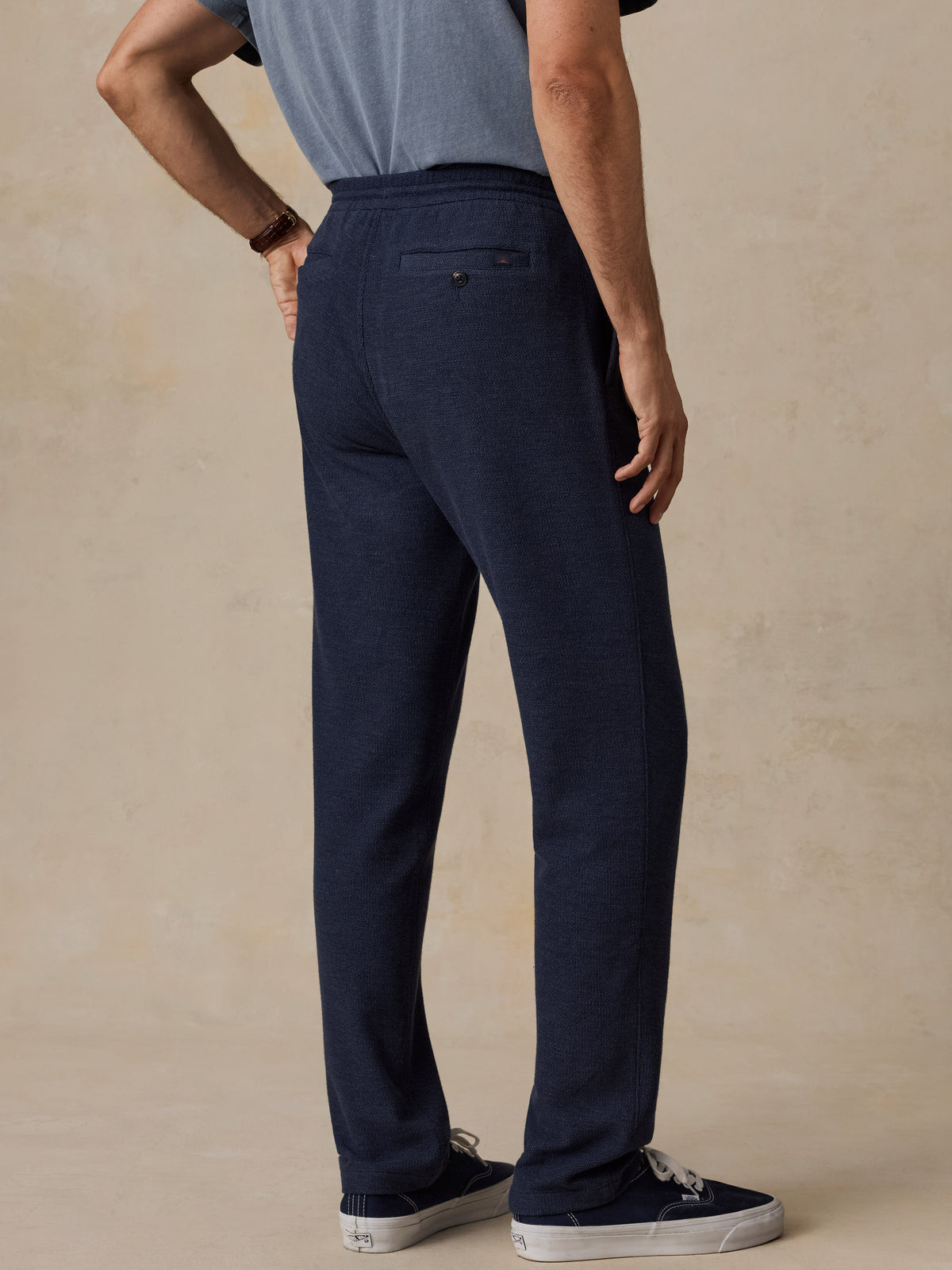 A man shown from the back wearing dark navy blue drawstring trousers, a light blue t-shirt, and navy sneakers against a textured beige studio backdrop., image 3 of 6