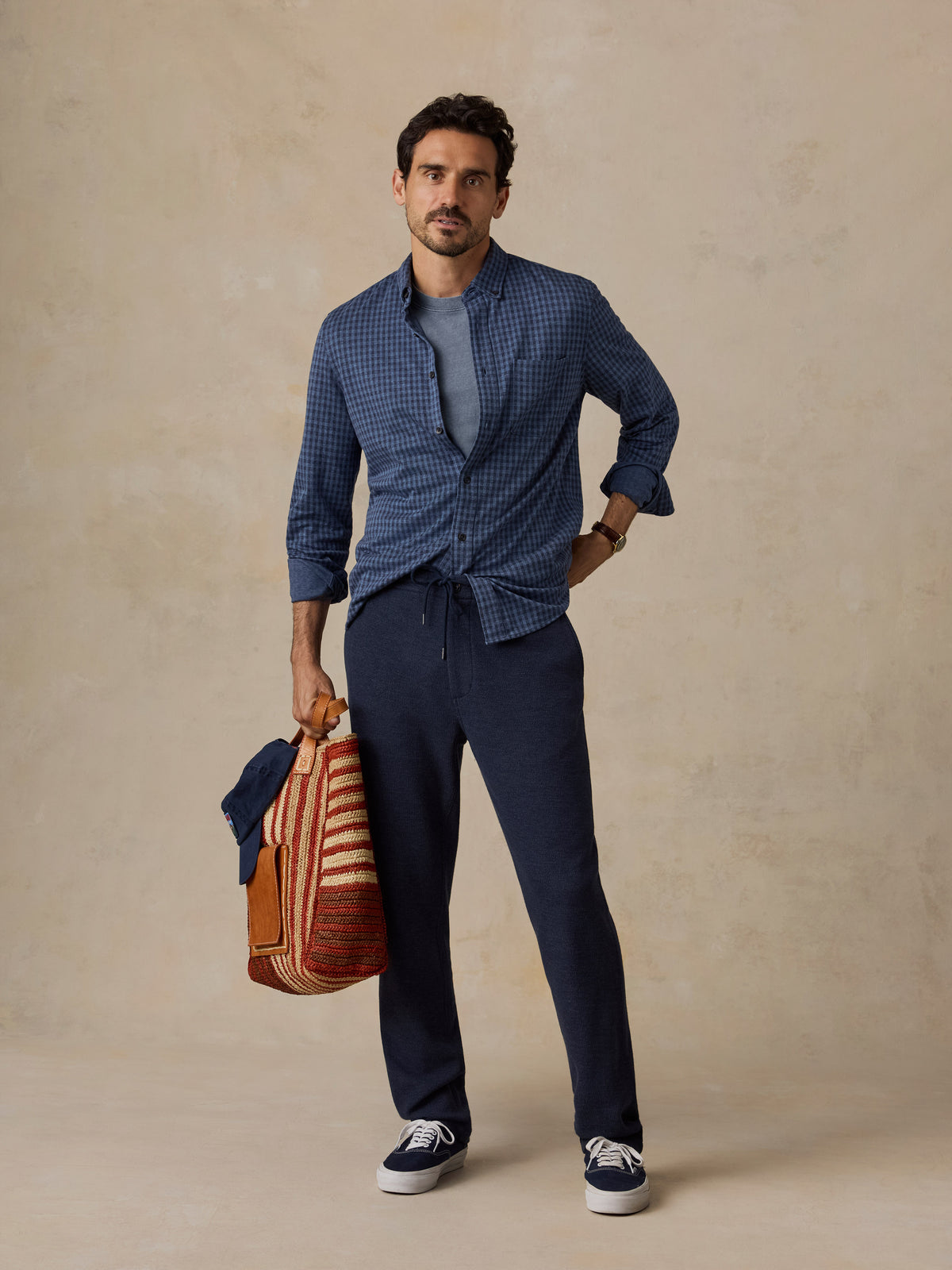 A man stands against a textured beige studio backdrop wearing a blue patterned button-up shirt tied at the waist over a gray T-shirt, navy drawstring pants, and navy sneakers. He is holding a striped brown and orange bag., image 5 of 6