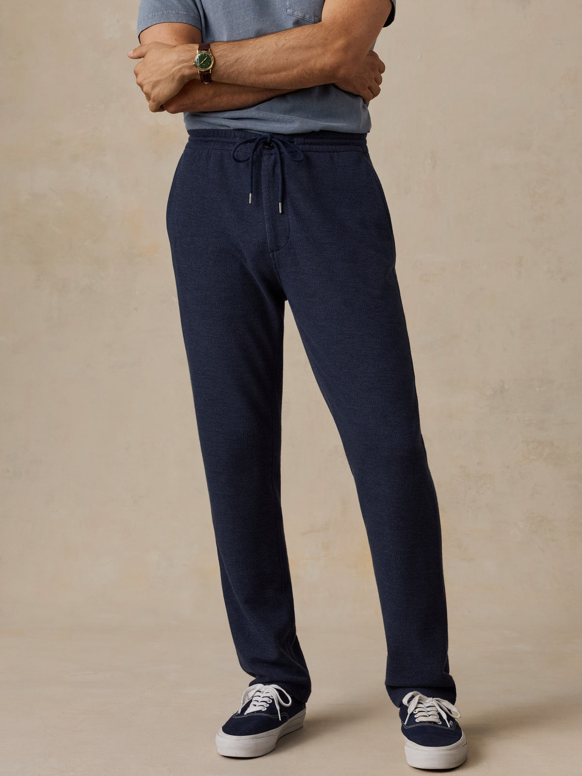 A man wearing dark navy drawstring sweatpants, a blue-gray t-shirt, and navy sneakers, standing against a textured beige studio background., image 1 of 6