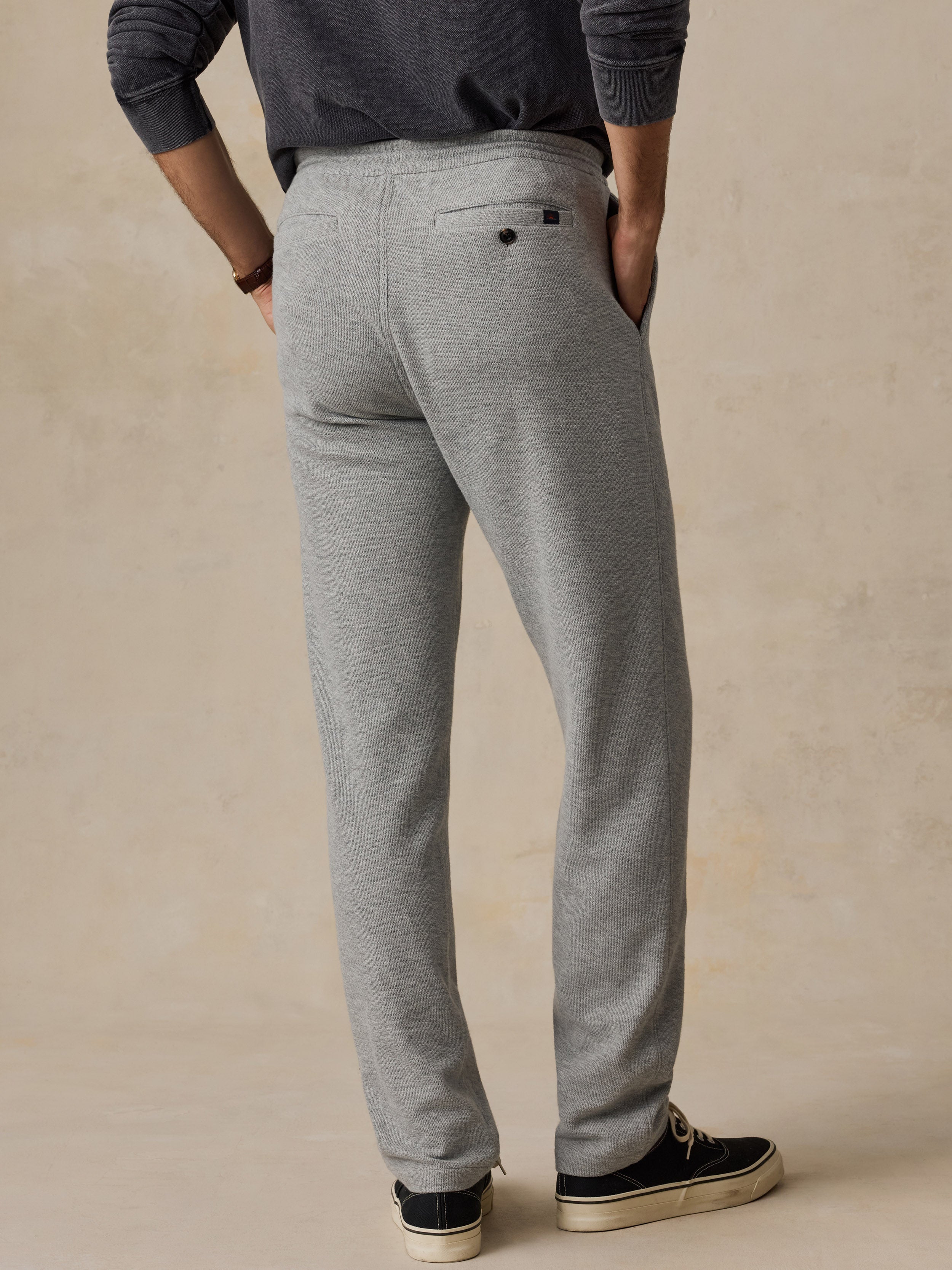 A man shown from the back wearing heather gray sweatpants, a dark gray long-sleeve shirt, and black sneakers against a warm beige textured studio background.