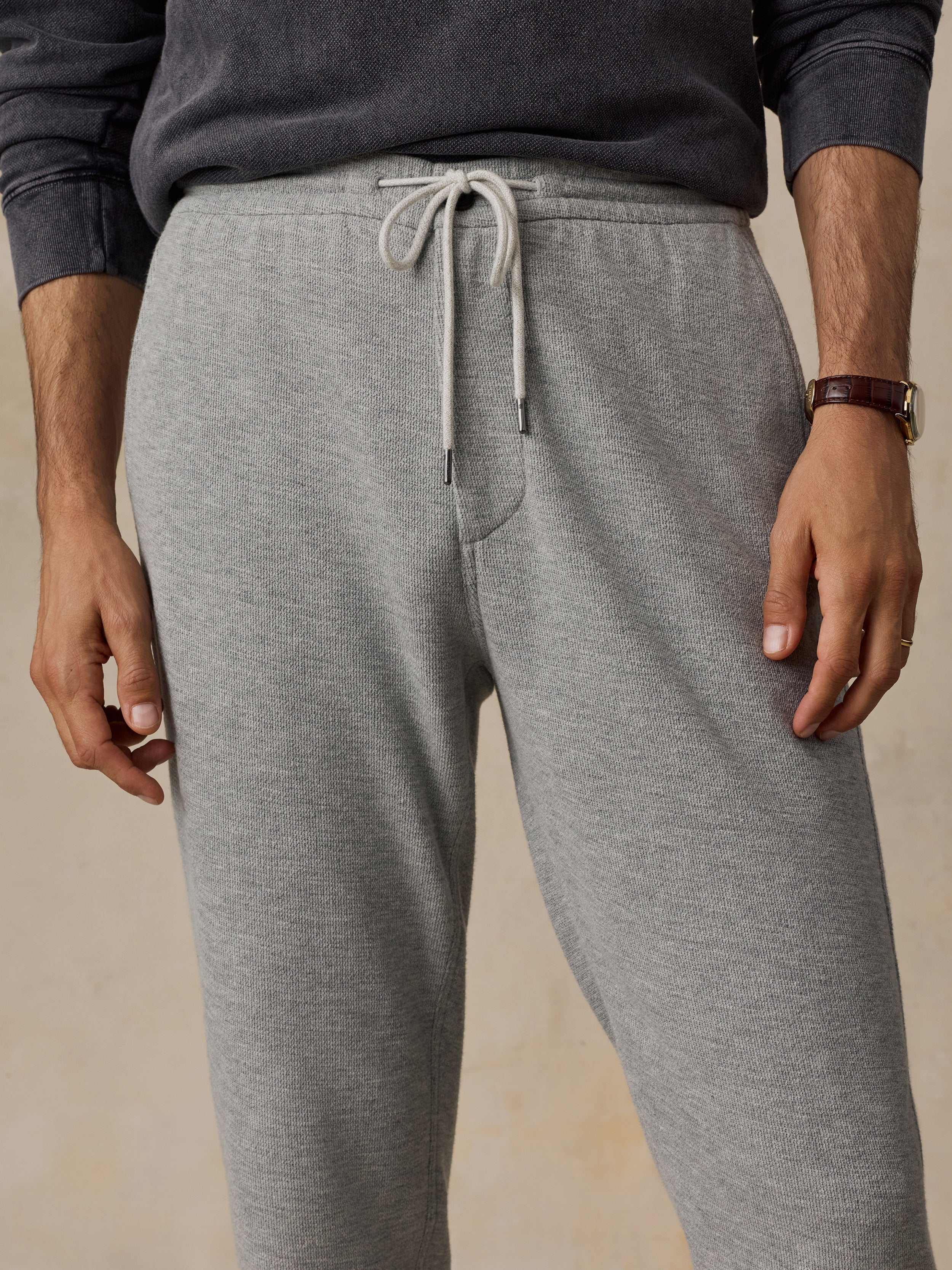 A close-up of a man wearing heather gray drawstring sweatpants and a dark charcoal long-sleeve top, standing against a warm beige studio background.