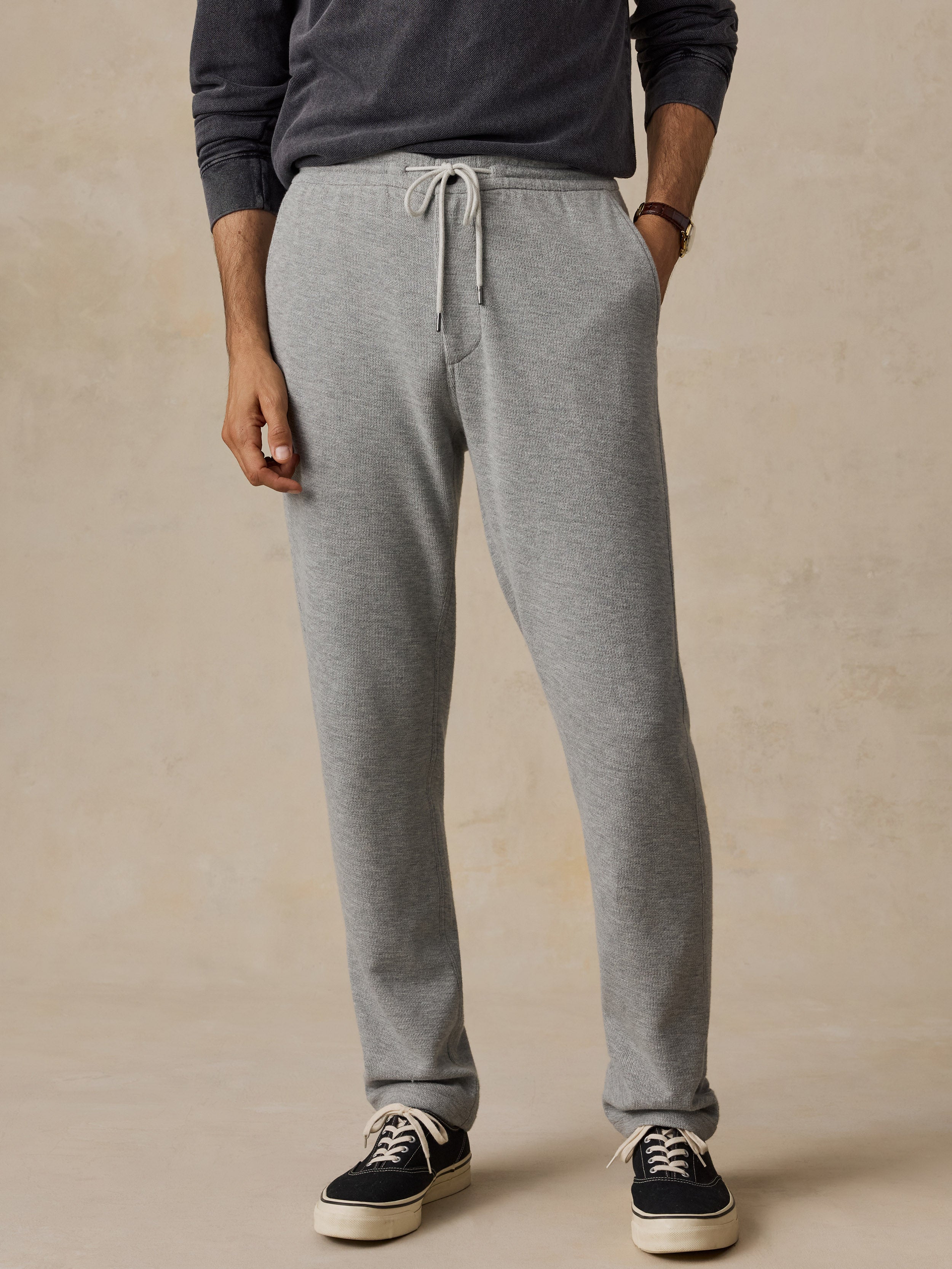 A man wearing light heather gray drawstring sweatpants, a dark gray long-sleeve top, and black low-top sneakers, standing against a warm beige textured studio background.