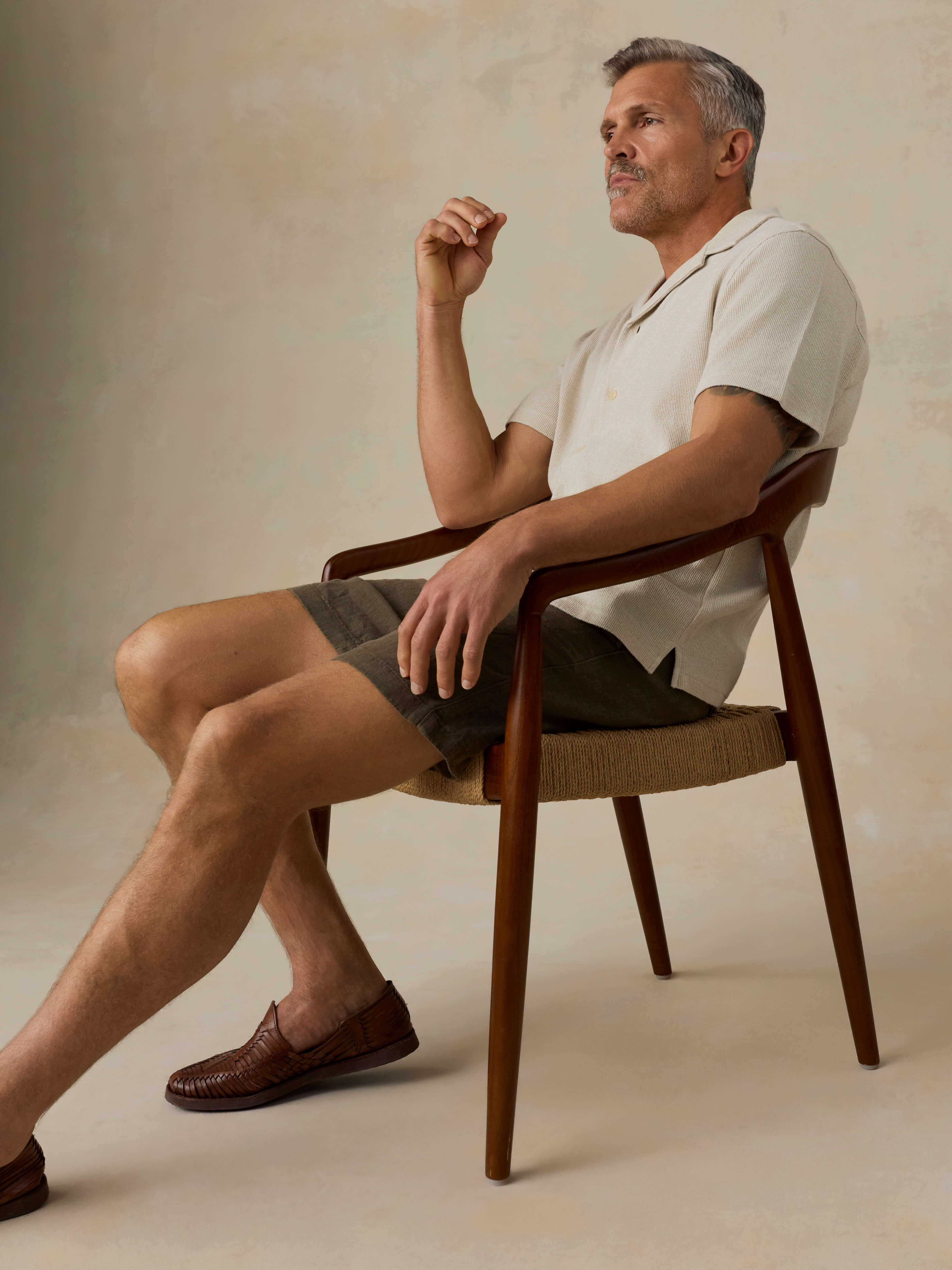 Man sitting on a chair wearing a light-colored shirt and green shorts against a beige background