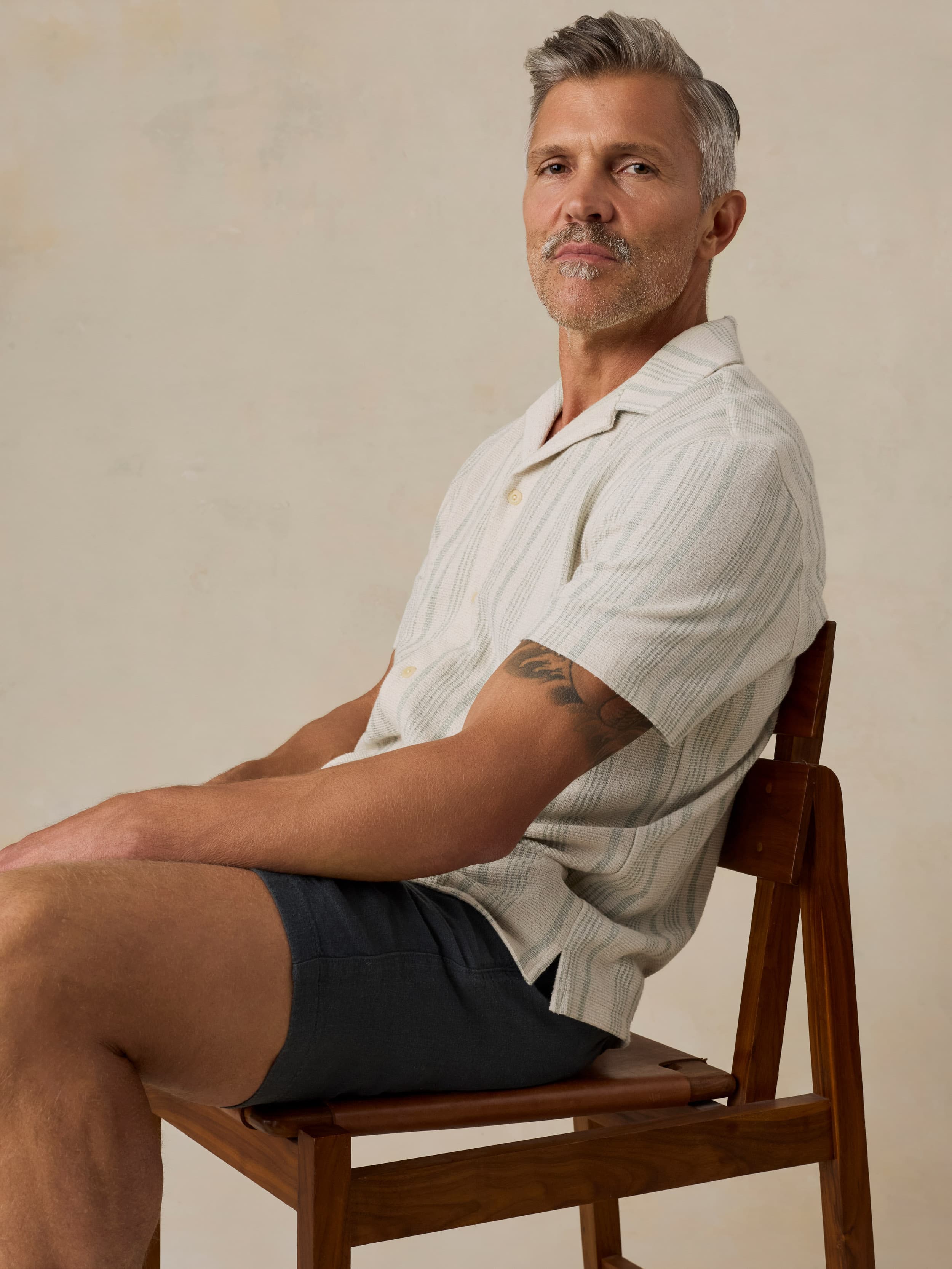Man sitting on a wooden chair wearing a light-colored shirt and black linen shorts against a beige background