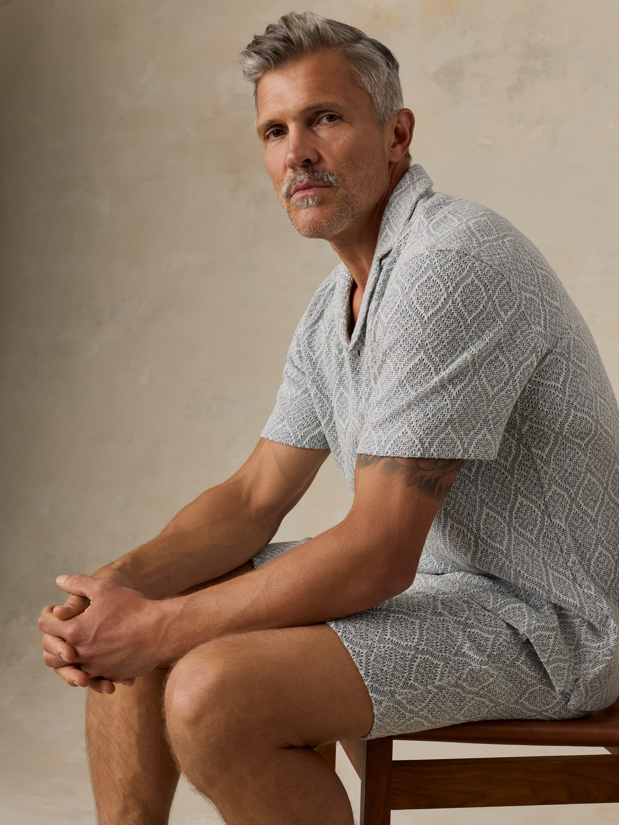 Man sitting on a wooden chair wearing blue printed shorts and a white t-shirt against a beige background