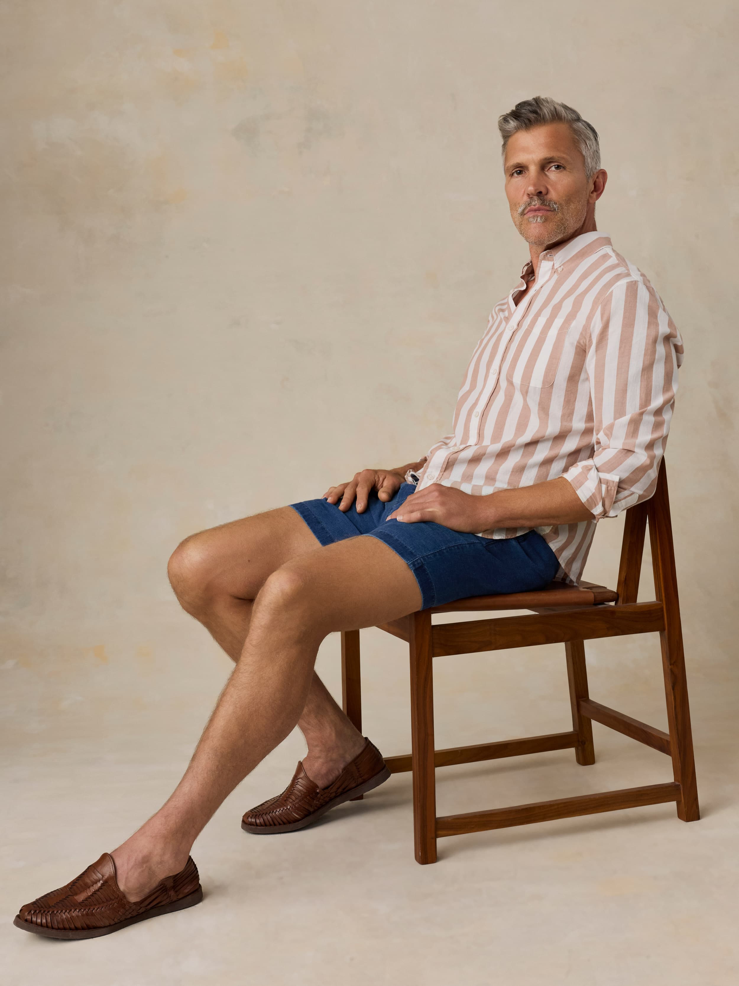 Man sitting on a wooden chair wearing a striped shirt and blue linen shorts against a beige background