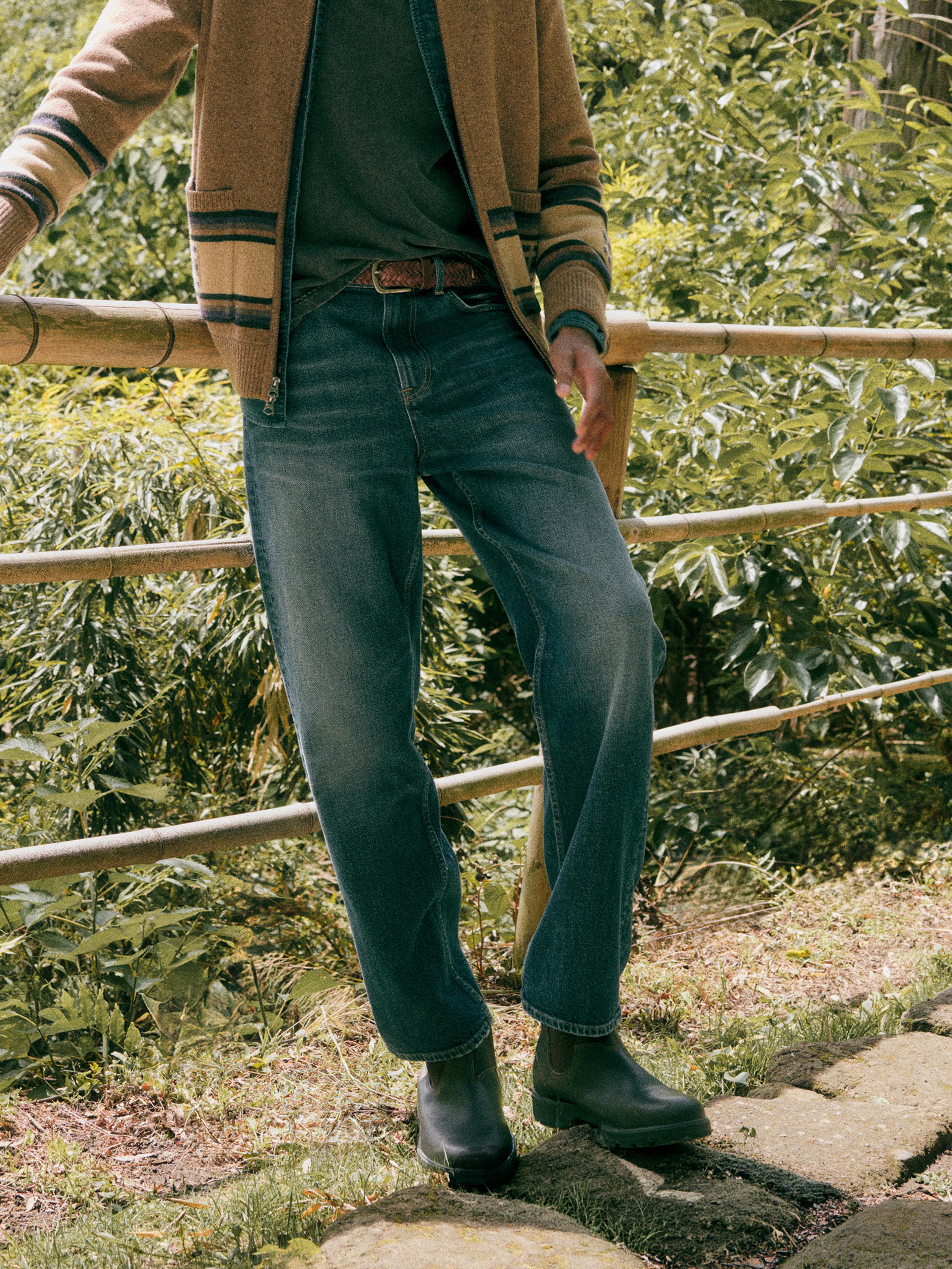 A man outdoors wearing blue denim jeans, a brown jacket, a dark green shirt, and dark rain boots, standing near a bamboo fence with green foliage in the background.
