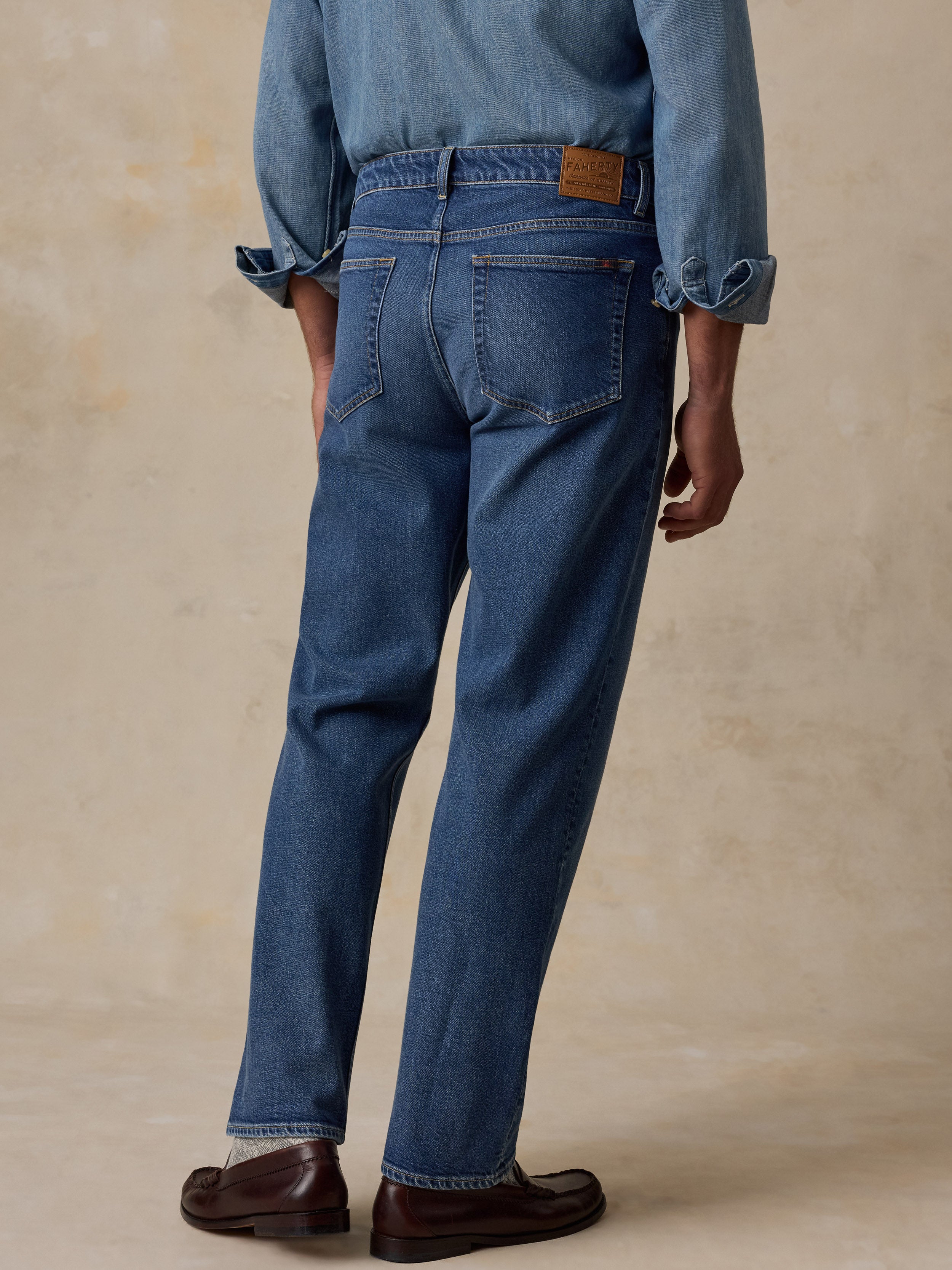 Rear view of a person wearing blue denim jeans with rolled-up sleeves on a light beige textured studio background.
