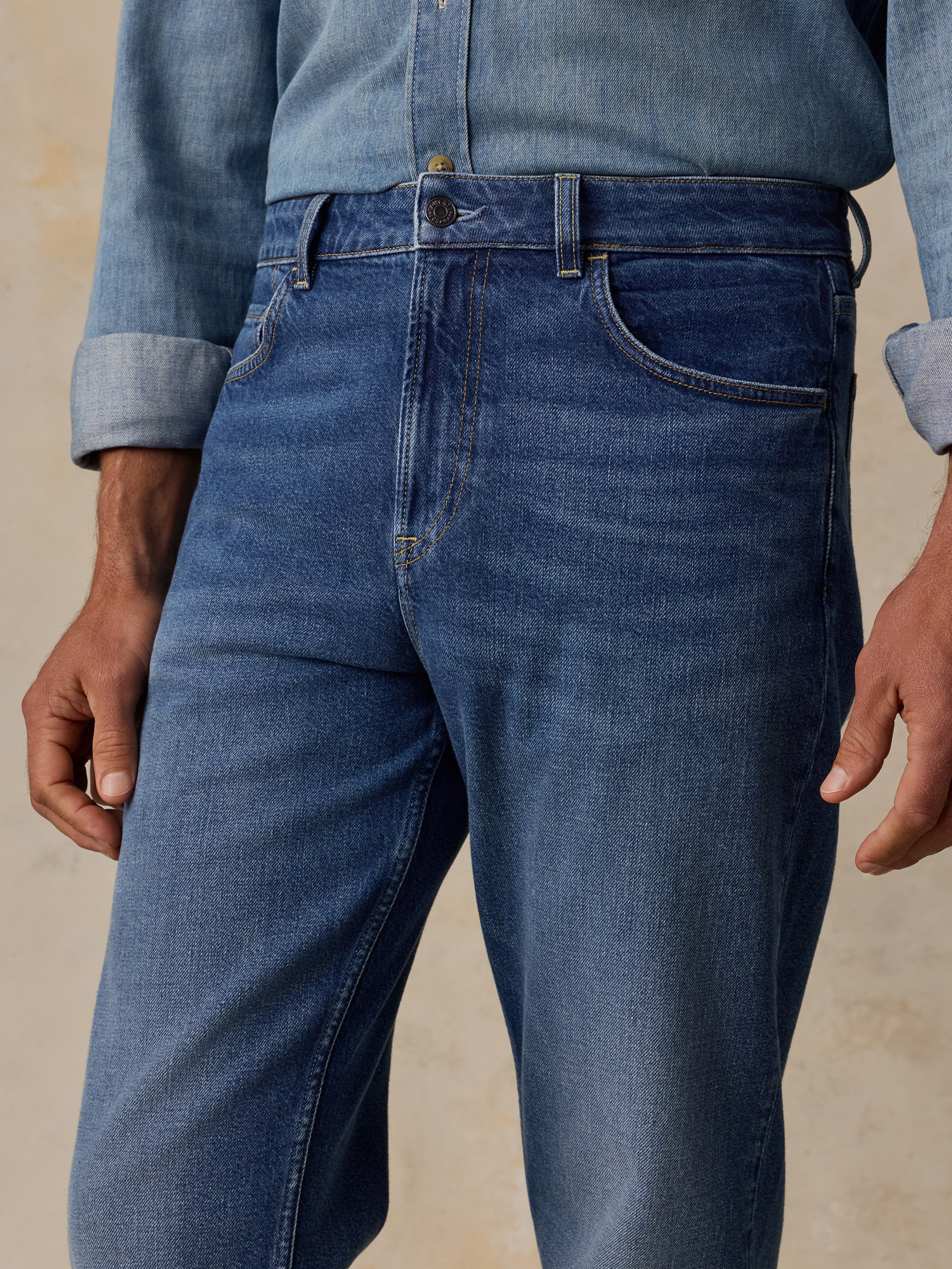 Close-up of a person wearing medium blue denim jeans with a rolled-up light blue denim shirt, shown from the waist to upper thighs against a warm beige textured background.
