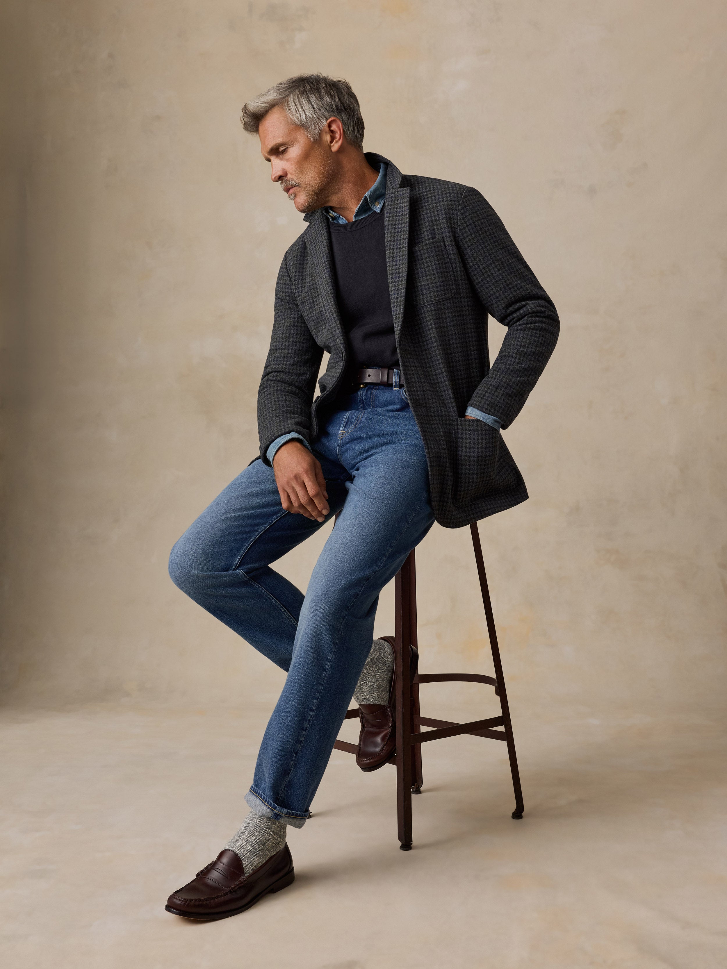 A man wearing a dark patterned blazer, blue jeans, a dark sweater, and brown loafers sits on a wooden stool against a textured beige studio backdrop.