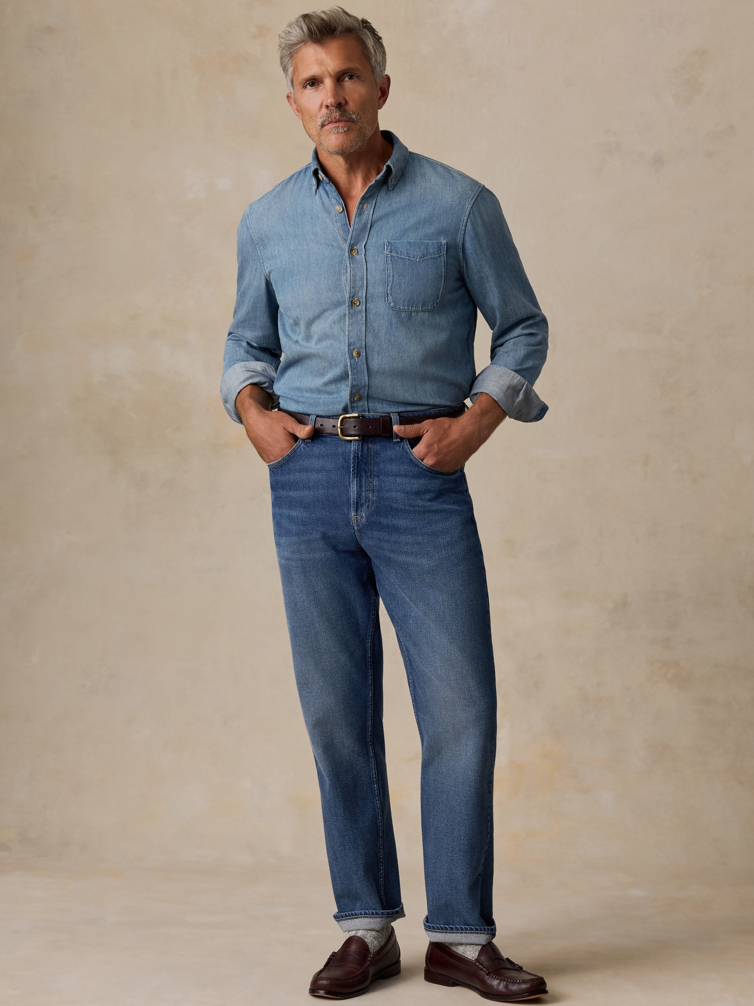 A man modeling a light blue denim button-down shirt and matching blue jeans, paired with a brown belt and dark brown leather loafers, standing against a textured beige studio backdrop.