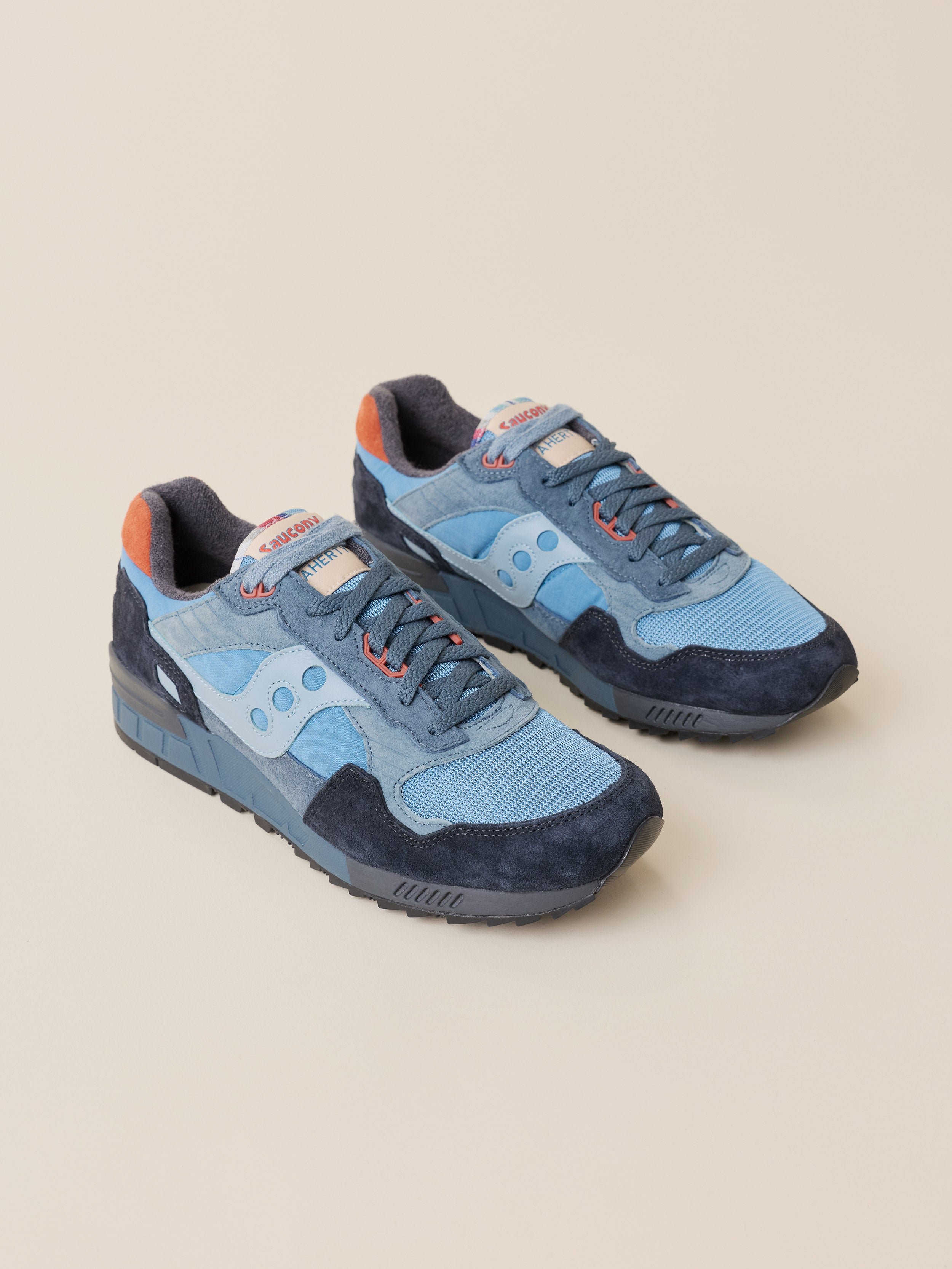 A pair of light blue and navy blue suede and mesh Faherty x Saucony sneakers with orange accents, displayed on a solid beige background.
