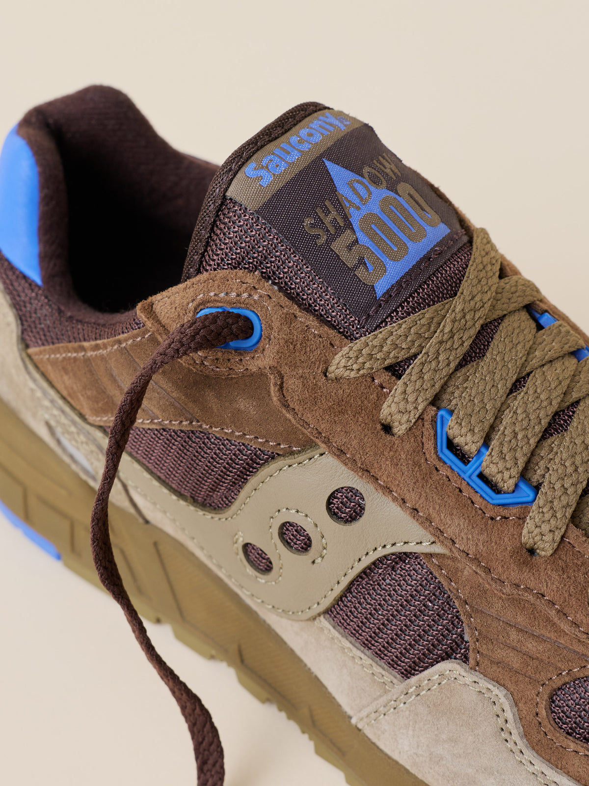 A close-up of a Saucony shoe in brown suede and mesh with beige overlays, olive laces, and bright blue accents on the tongue and lace loops., image 3 of 4