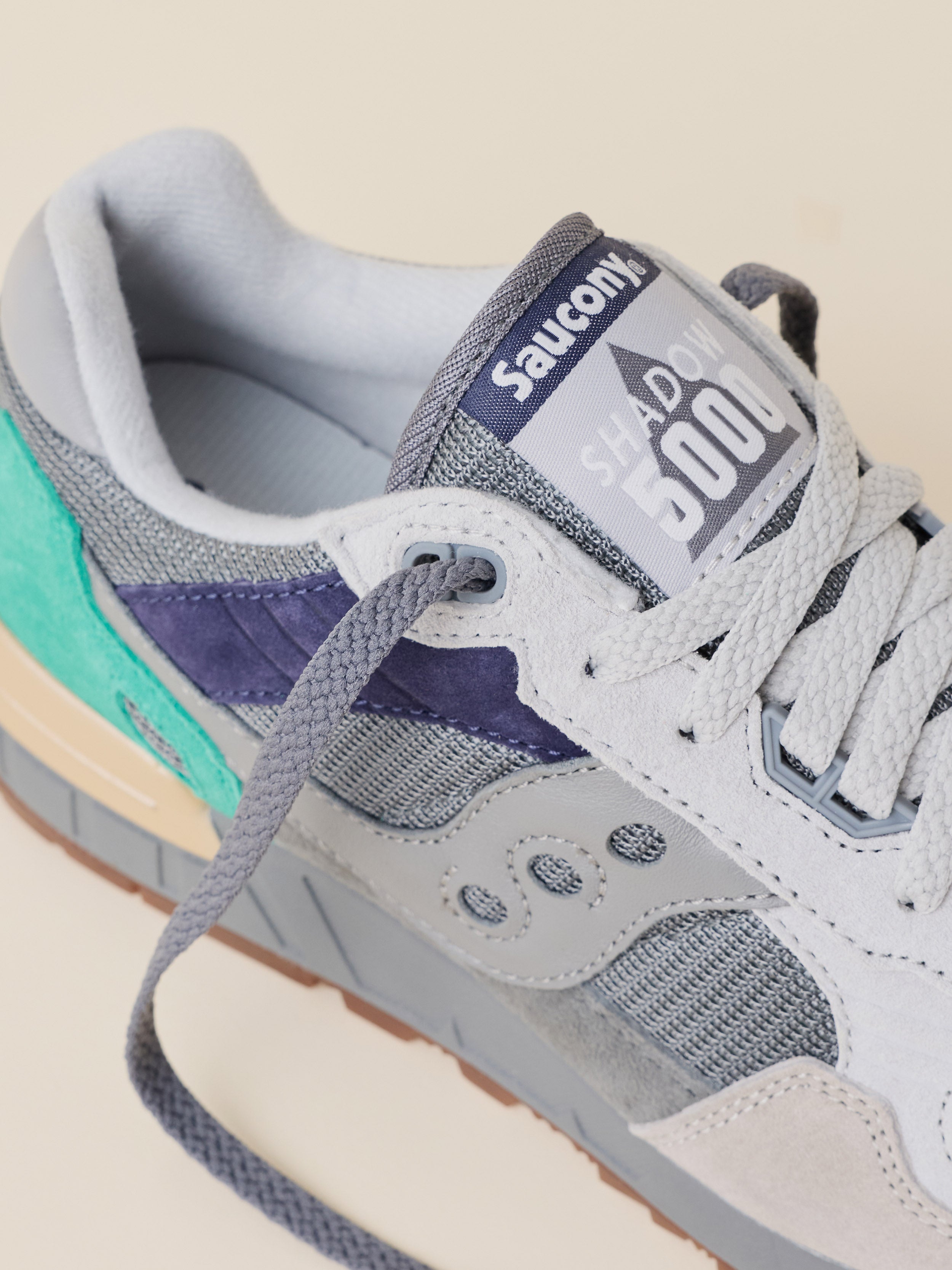 Close-up of a Saucony Shadow 500 sneaker in light gray suede and mesh with purple overlays, a teal heel accent, and gray laces on a light beige background.