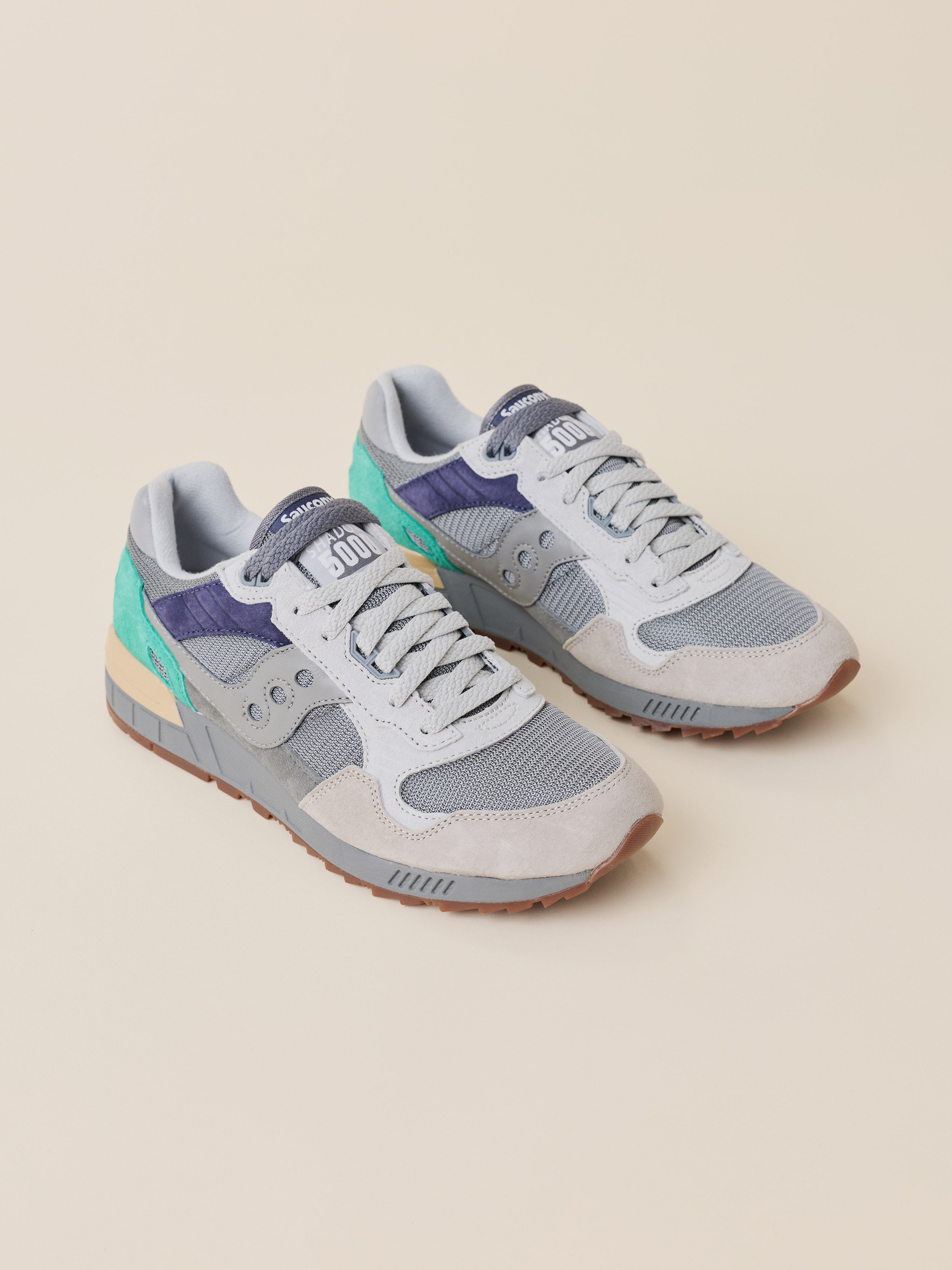 A pair of light gray and white Saucony sneakers with purple and mint green accents, displayed on a solid beige background.