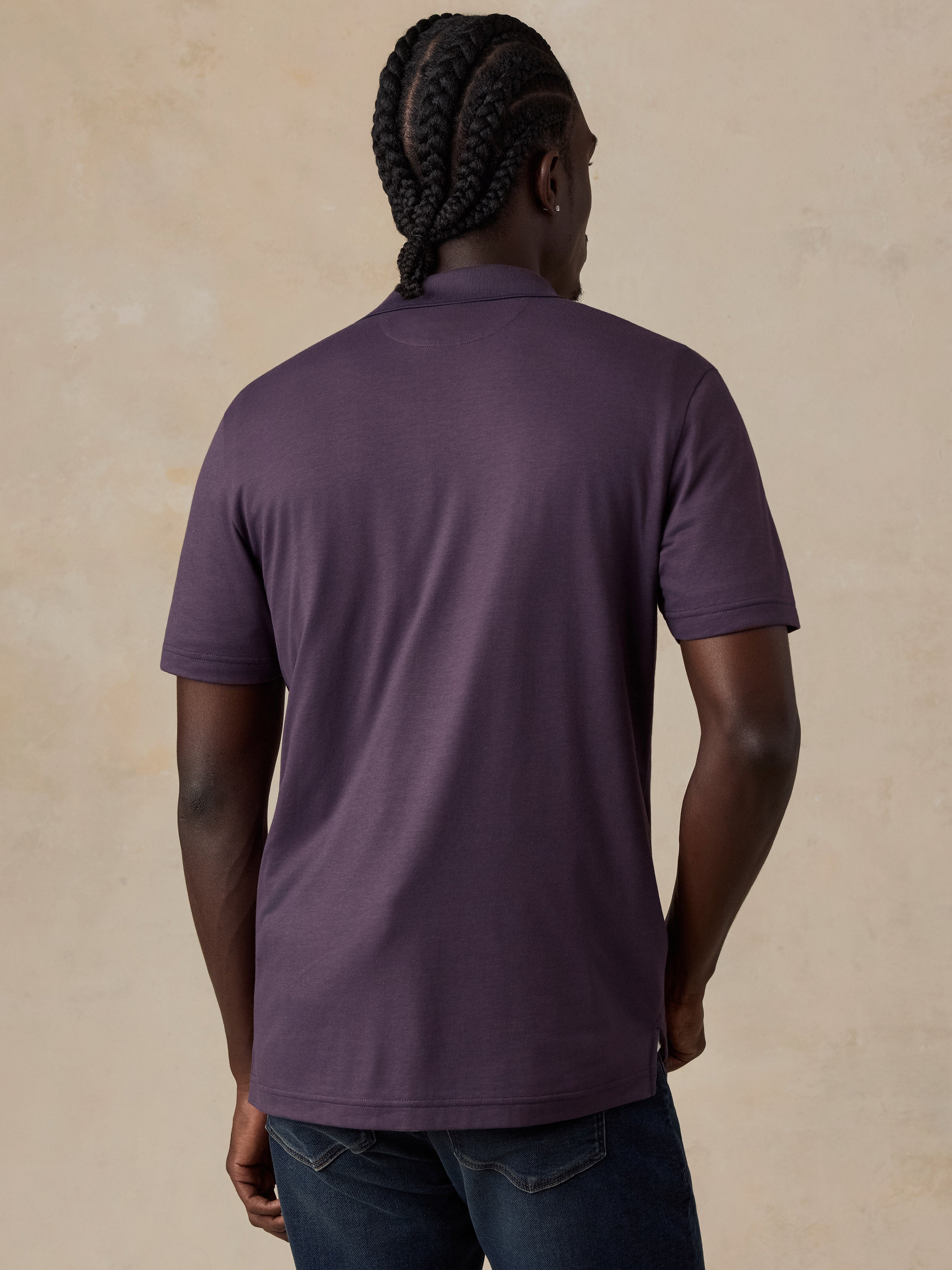 A man viewed from behind wearing a short-sleeve polo shirt in a muted plum purple color, paired with dark blue jeans, against a textured beige background.