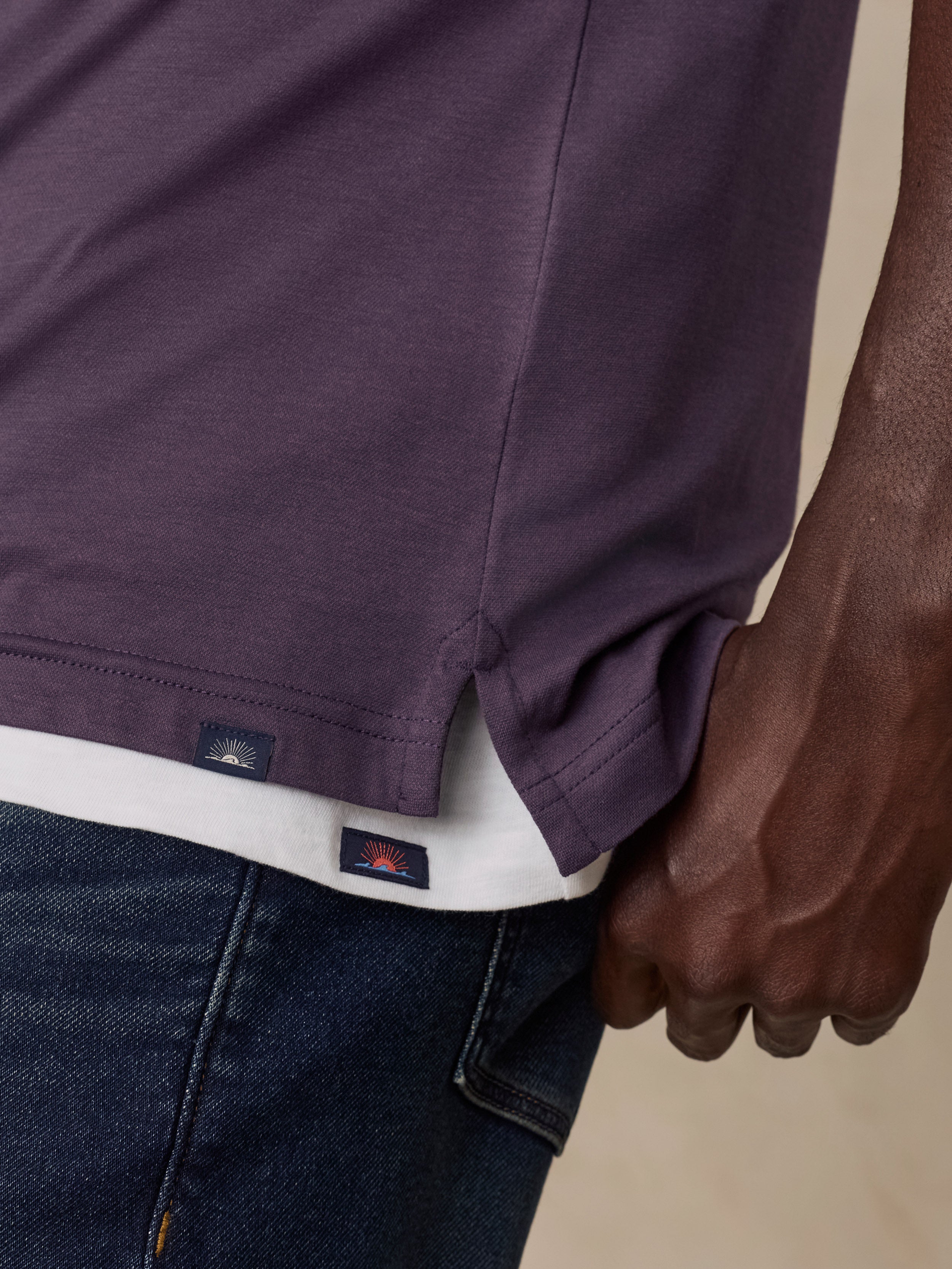 Close-up of a man wearing a dark purple t-shirt with a white undershirt and dark blue jeans, showing the side hem and Faherty brand tag.
