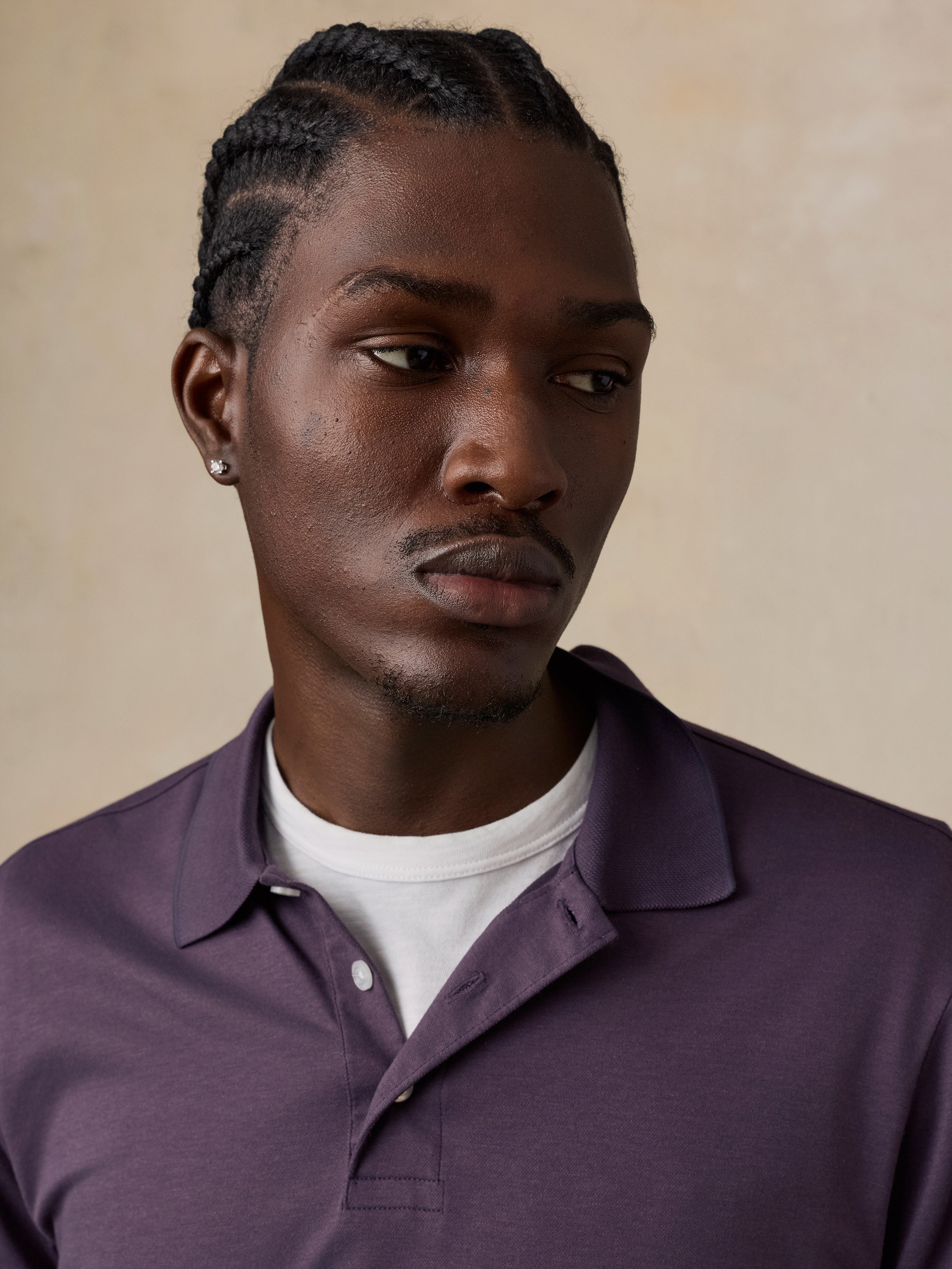 A close-up portrait of a man wearing a dark purple polo shirt over a white t-shirt, set against a warm beige textured background.