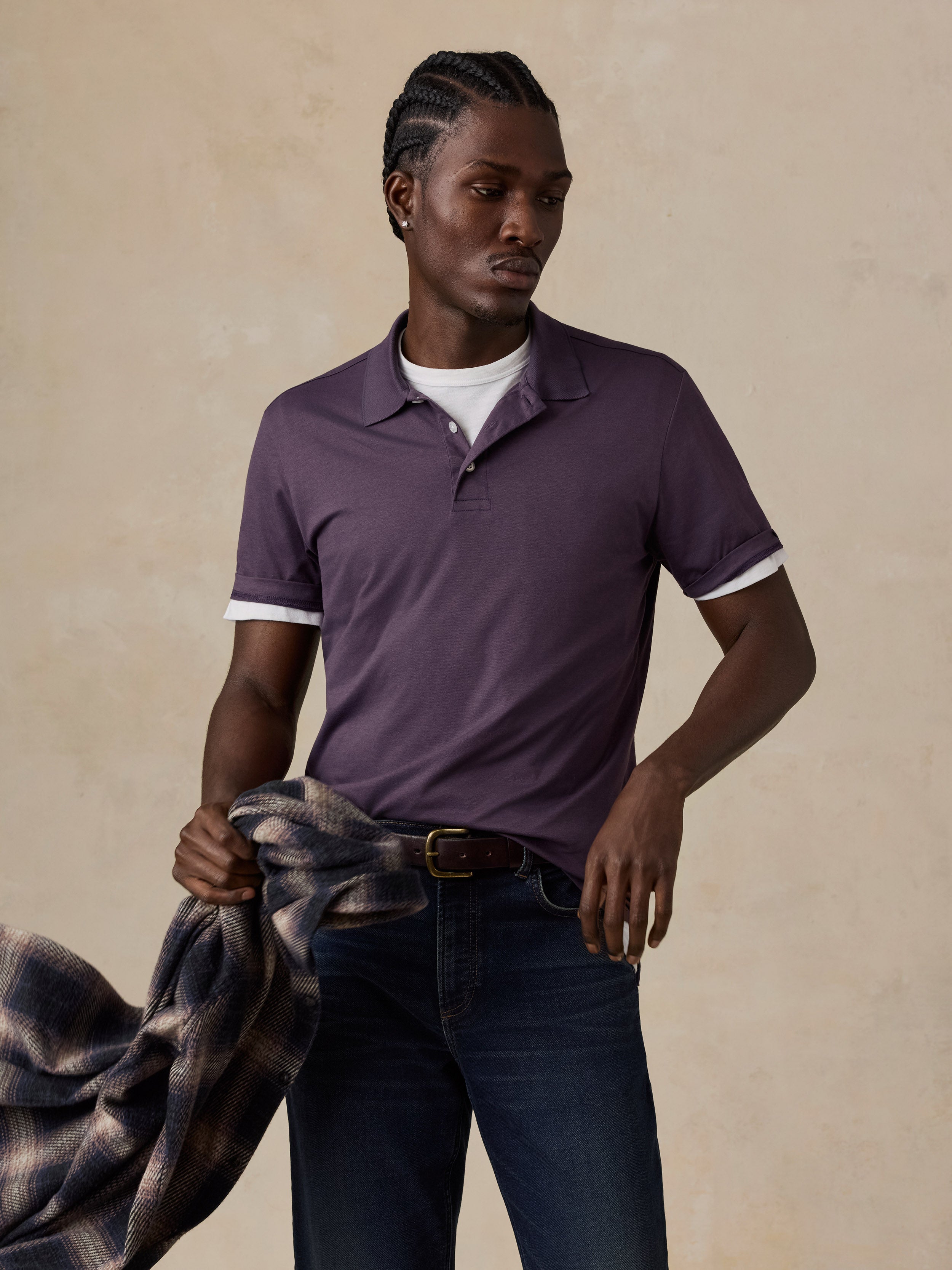 A man wearing a short-sleeve polo shirt in a muted plum purple, layered over a white crewneck t-shirt and paired with dark blue jeans. He is holding a plaid shirt in his hand, posed against a textured beige studio backdrop.