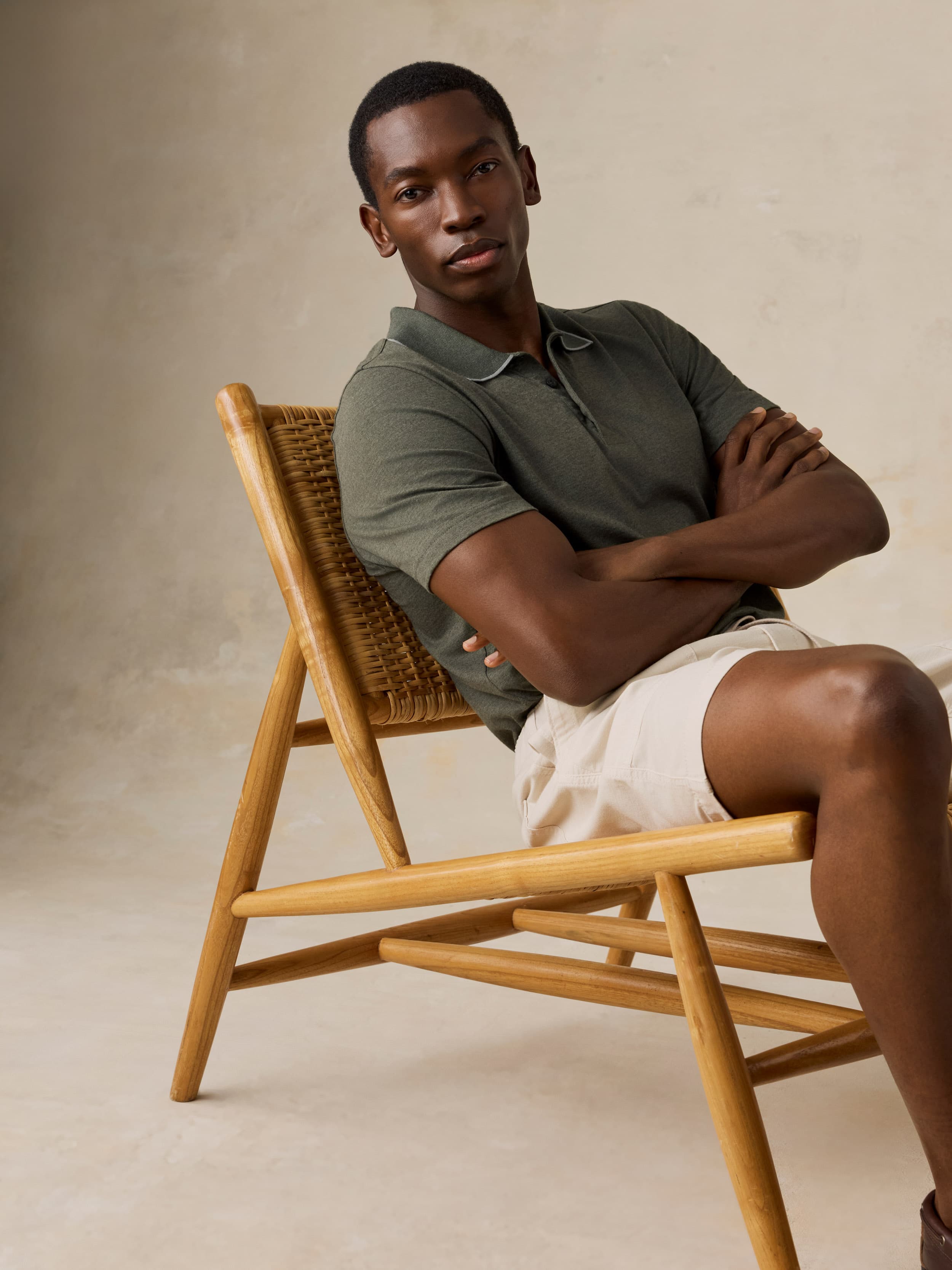 Man sitting on a wooden chair wearing a green polo and beige shorts against a beige background