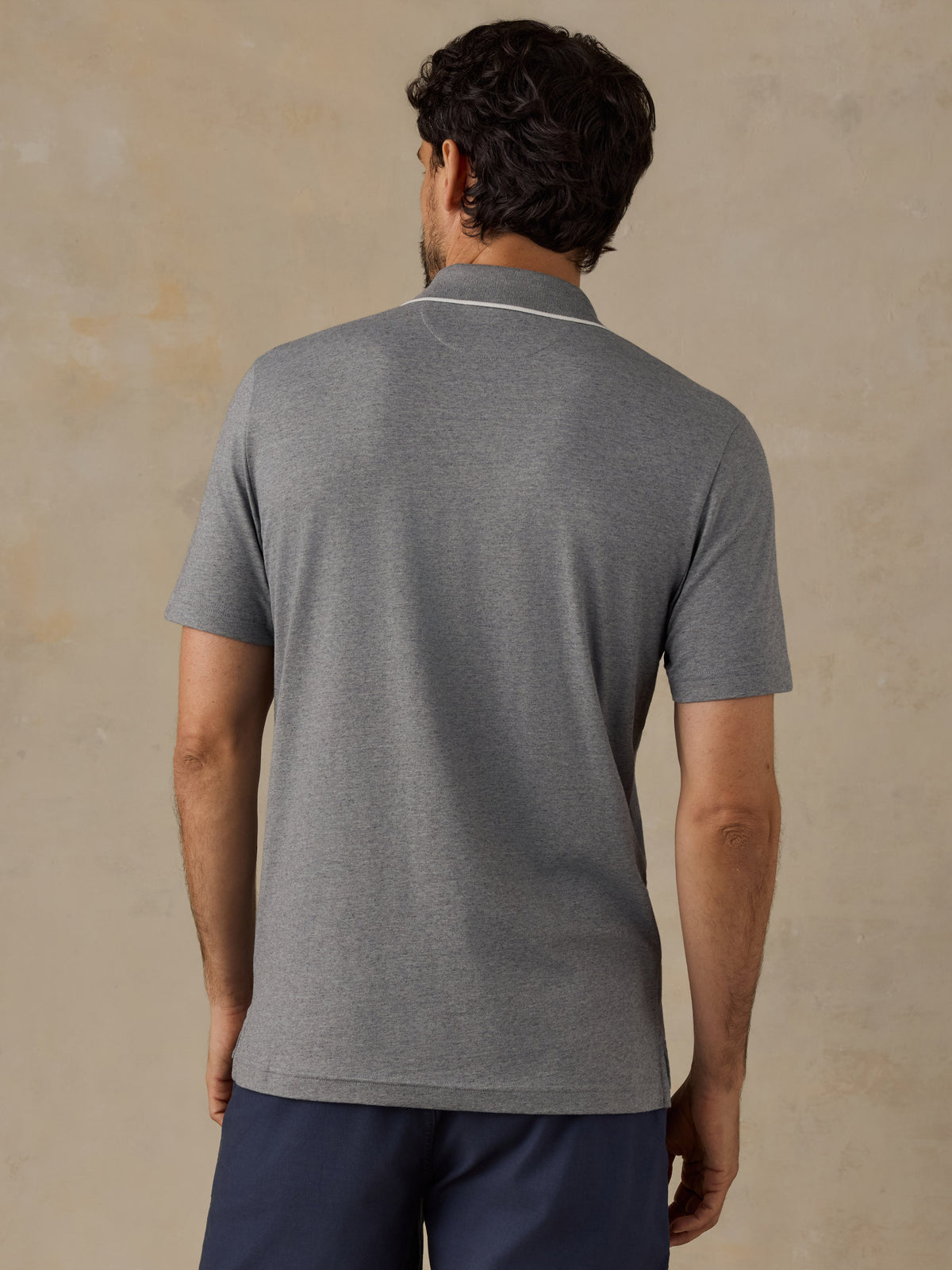 A man viewed from behind wearing a heather gray short-sleeve polo shirt with a thin white stripe detail at the collar, paired with navy blue pants, set against a textured beige background., image 3 of 6