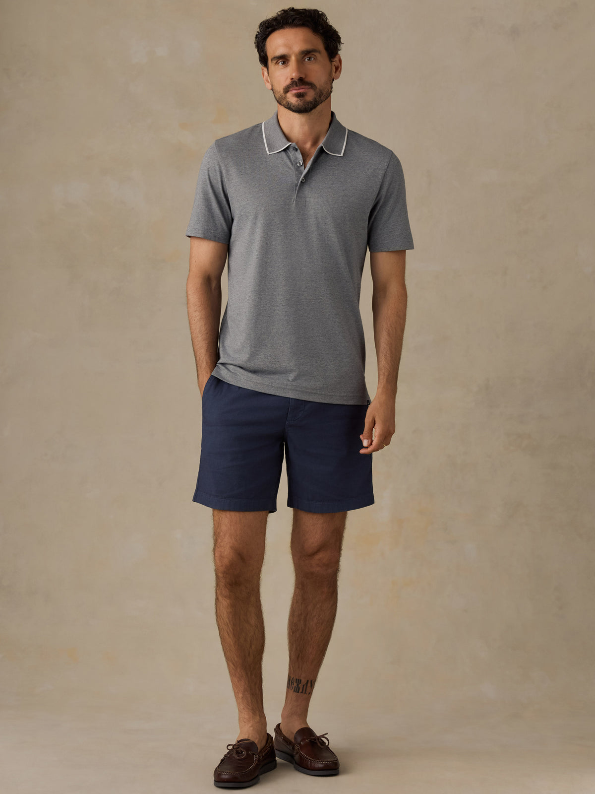A man modeling a short-sleeve polo shirt in heather gray with a white contrast collar, paired with navy blue shorts and brown boat shoes, standing against a textured beige studio backdrop., image 4 of 6