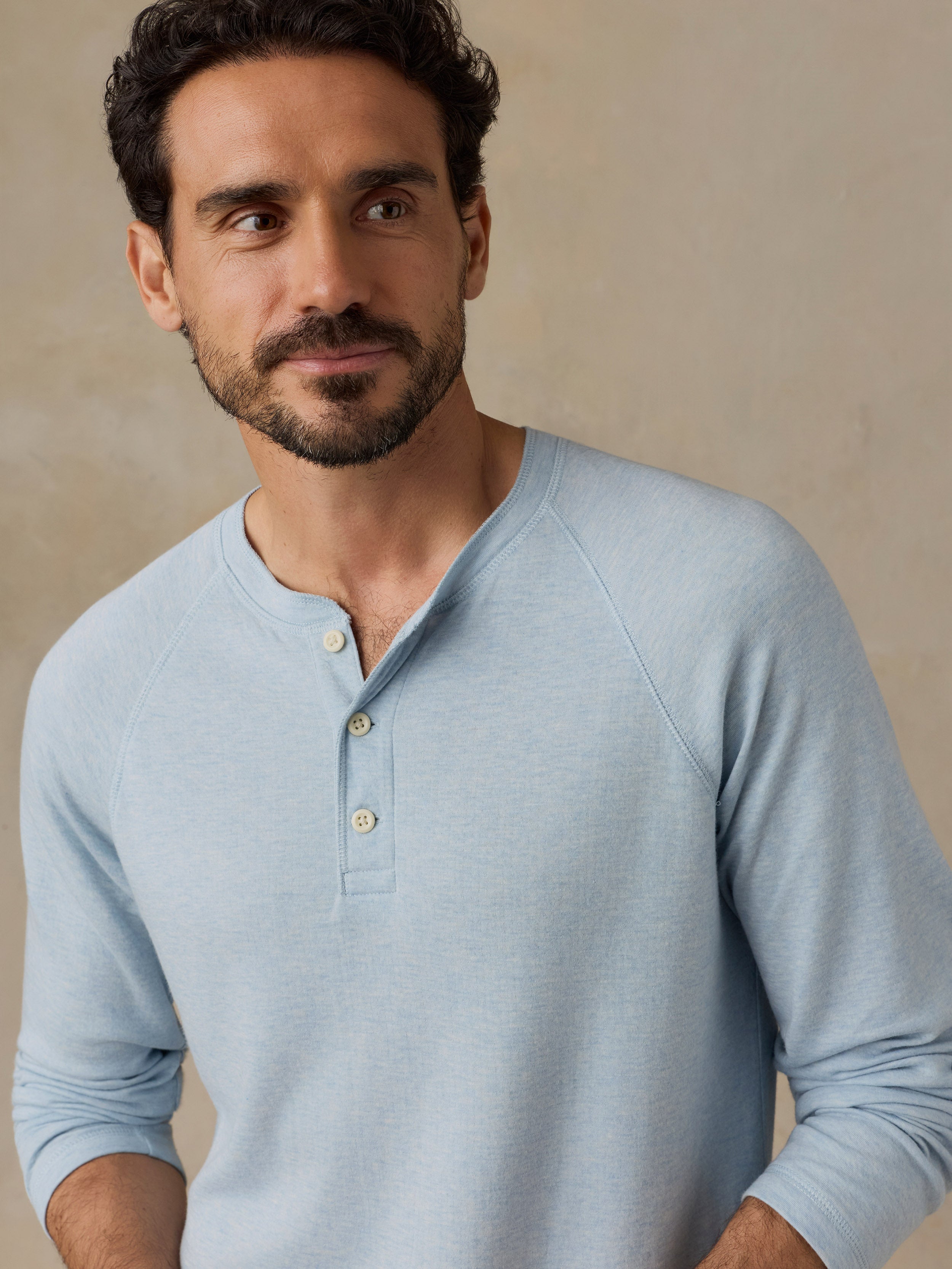 A man wearing a light blue long-sleeve henley shirt with three buttons, photographed against a neutral beige background.