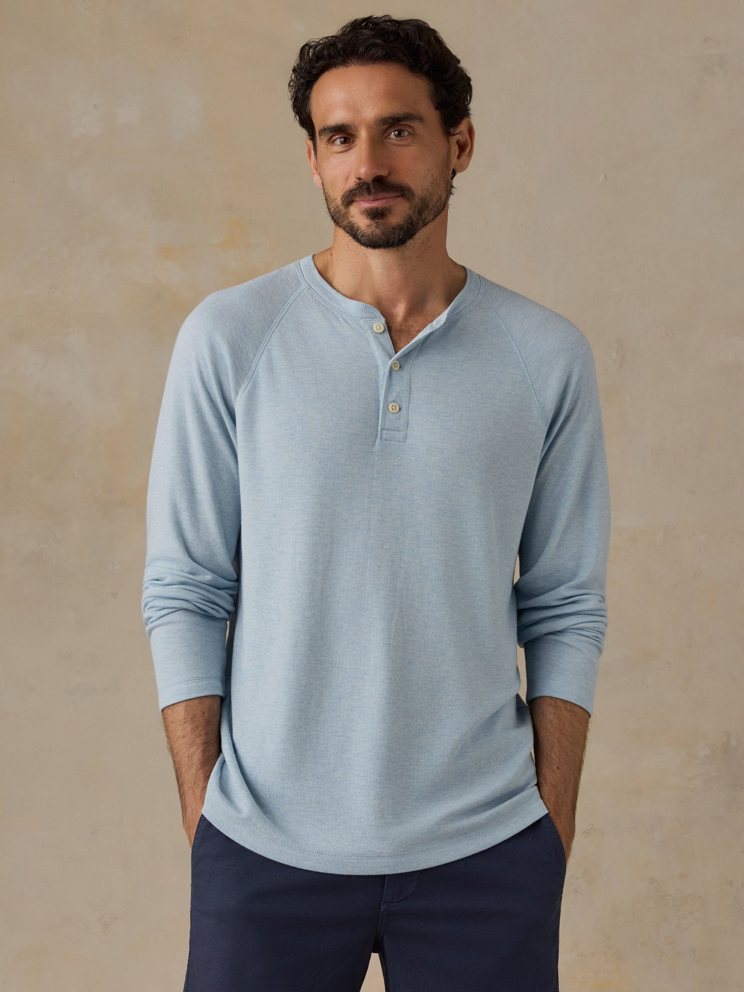 A man wearing a light blue long-sleeve henley shirt and navy pants, standing against a textured beige background.