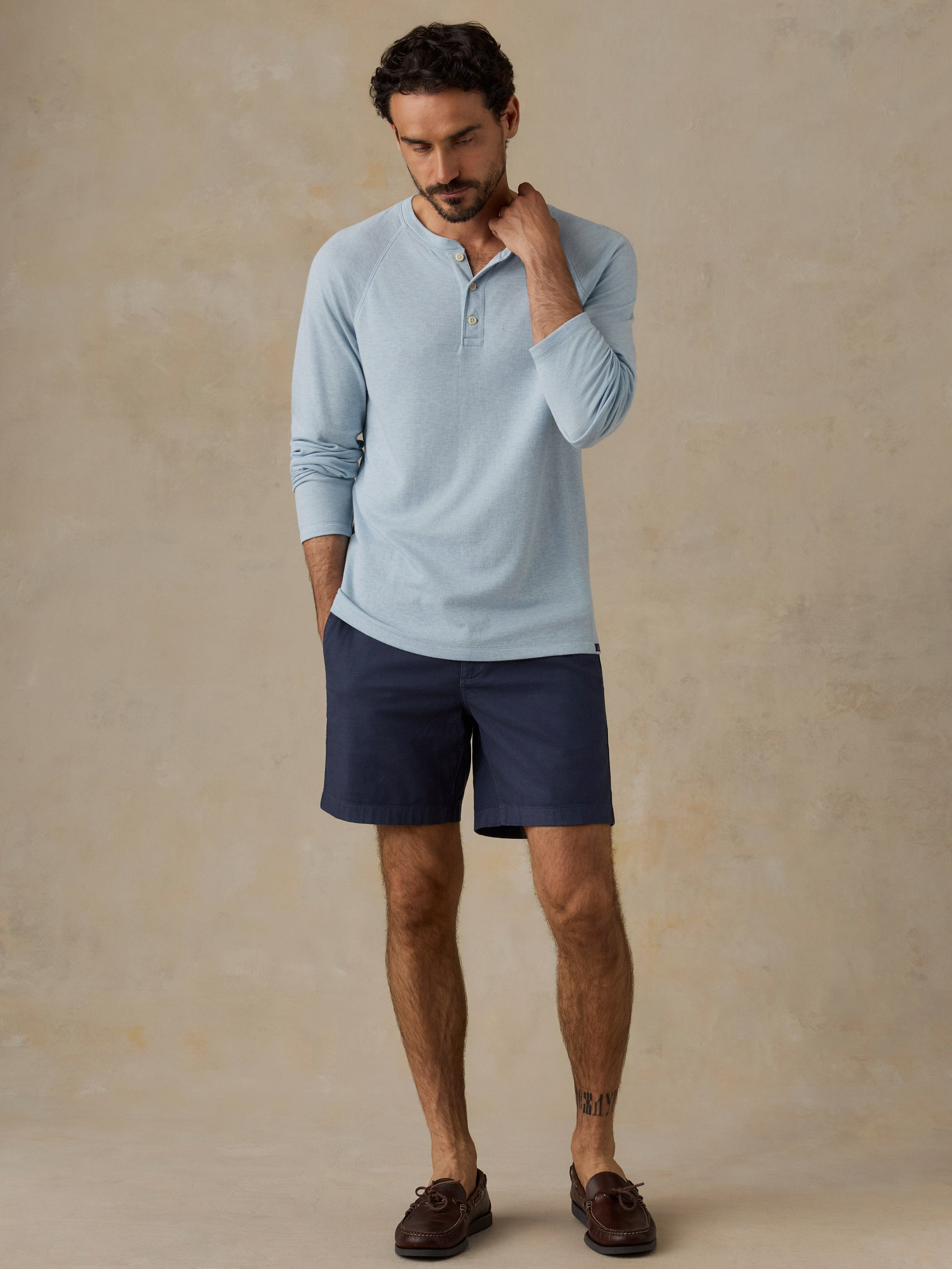 A man modeling a light blue long-sleeve henley shirt and navy blue shorts against a textured beige studio backdrop.