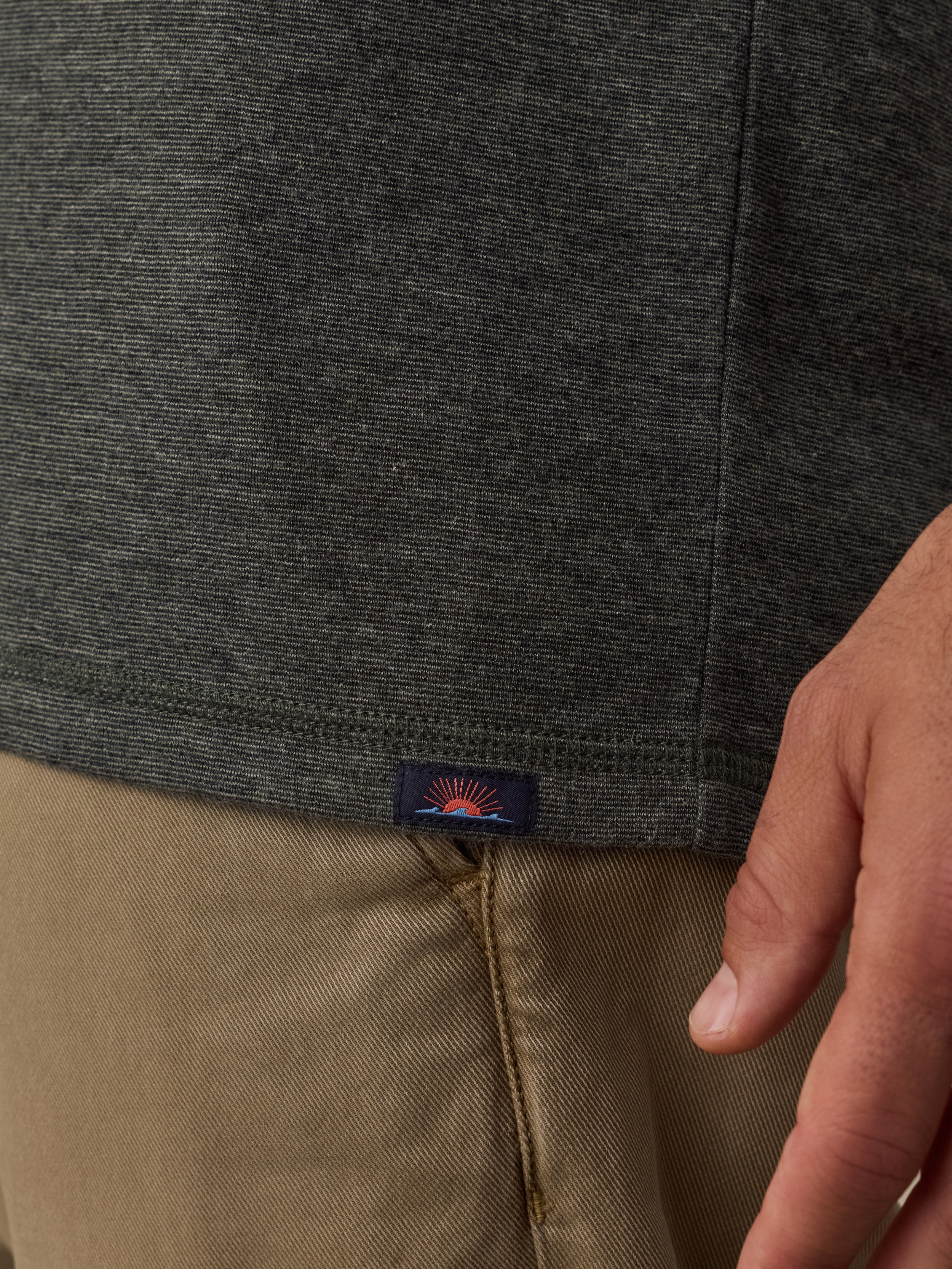 Close-up of a person wearing a dark green knit top and tan pants, with a small embroidered logo patch visible near the hem of the shirt.