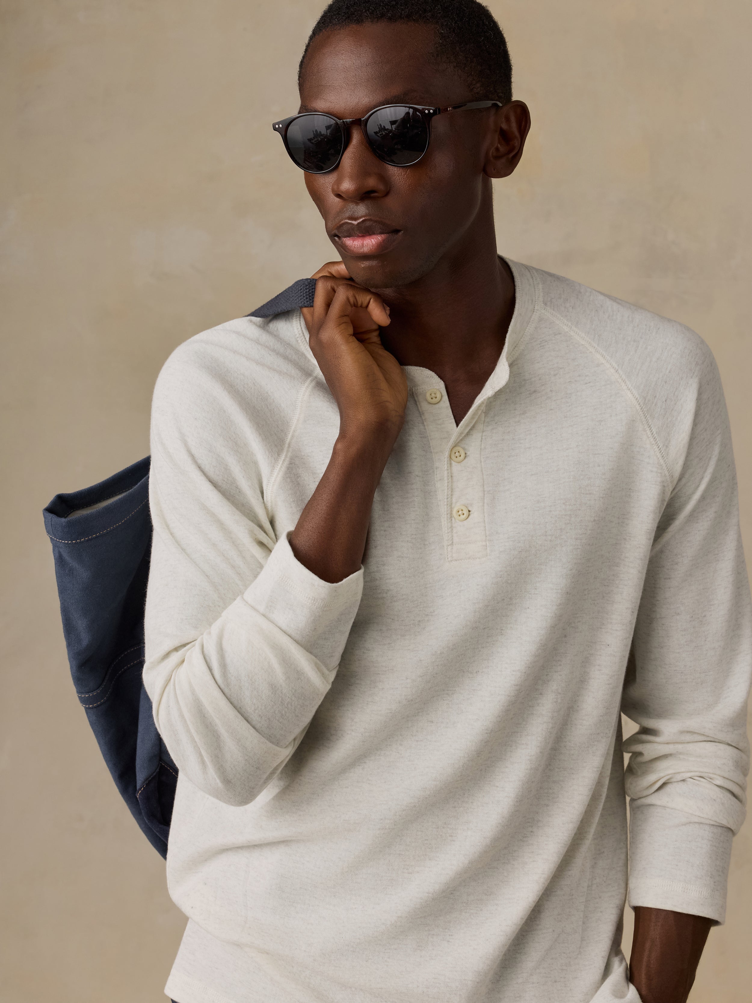 Man is wearing a cream colored long-sleeve henley shirt and navy tote bag against a beige background