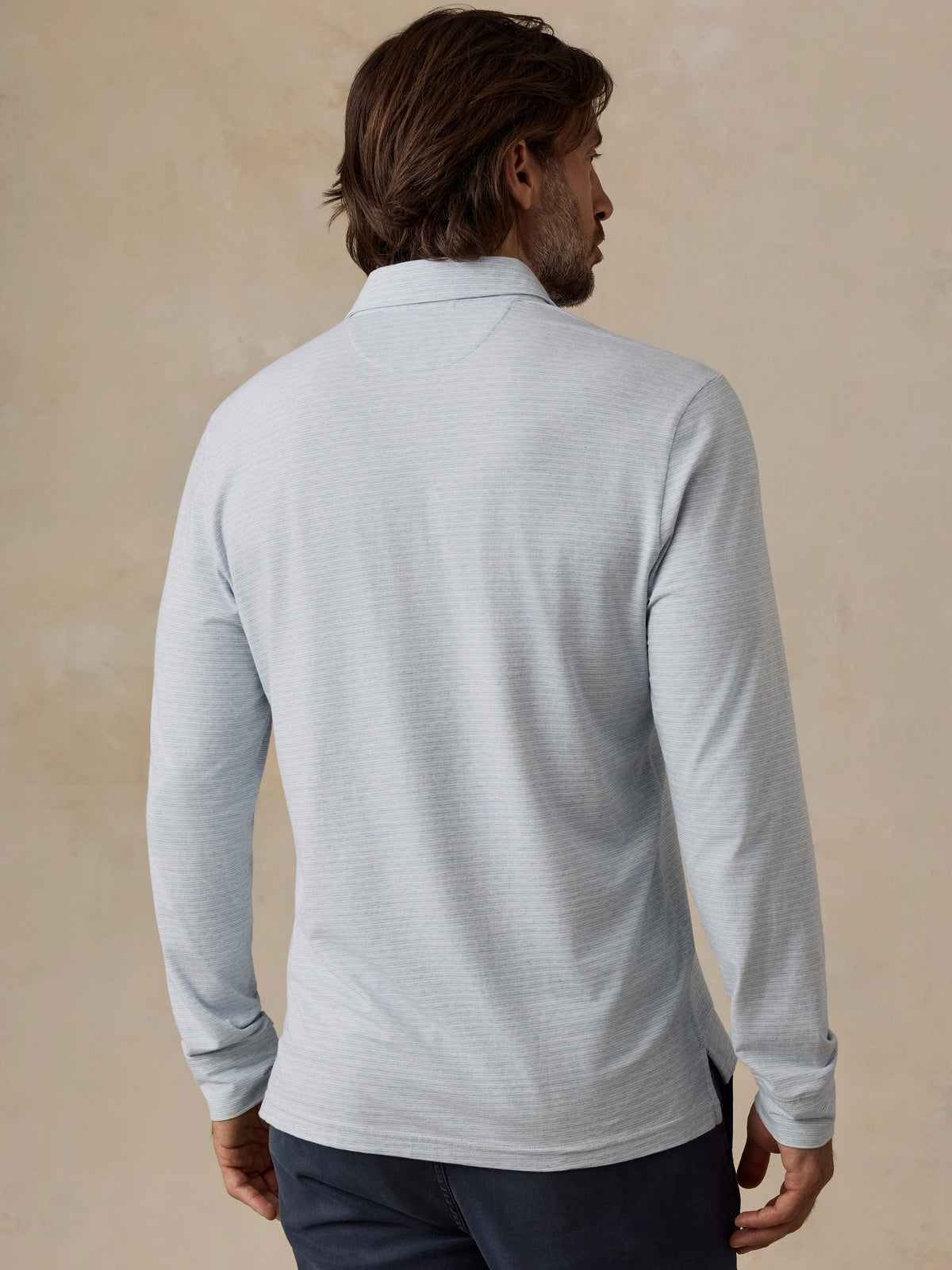 A man viewed from the back wearing a light blue long-sleeve polo shirt with a subtle textured stripe pattern, paired with dark navy pants, against a neutral beige textured wall., image 4 of 6