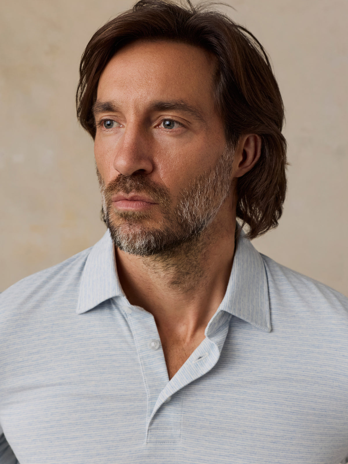 A close-up portrait of a man wearing a light blue and white striped polo shirt against a neutral beige background., image 3 of 6