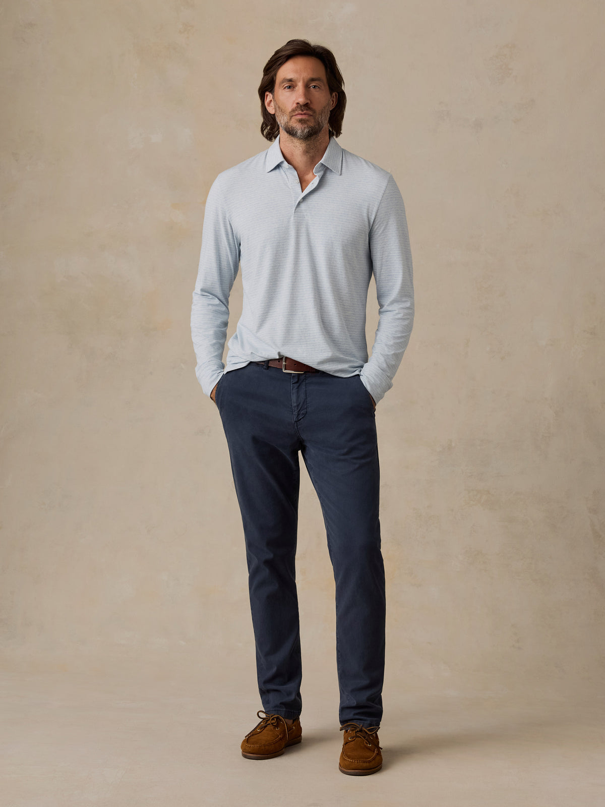 A man stands against a textured beige studio backdrop wearing a light blue long-sleeve polo shirt, navy blue trousers, a brown belt, and brown lace-up shoes., image 2 of 6