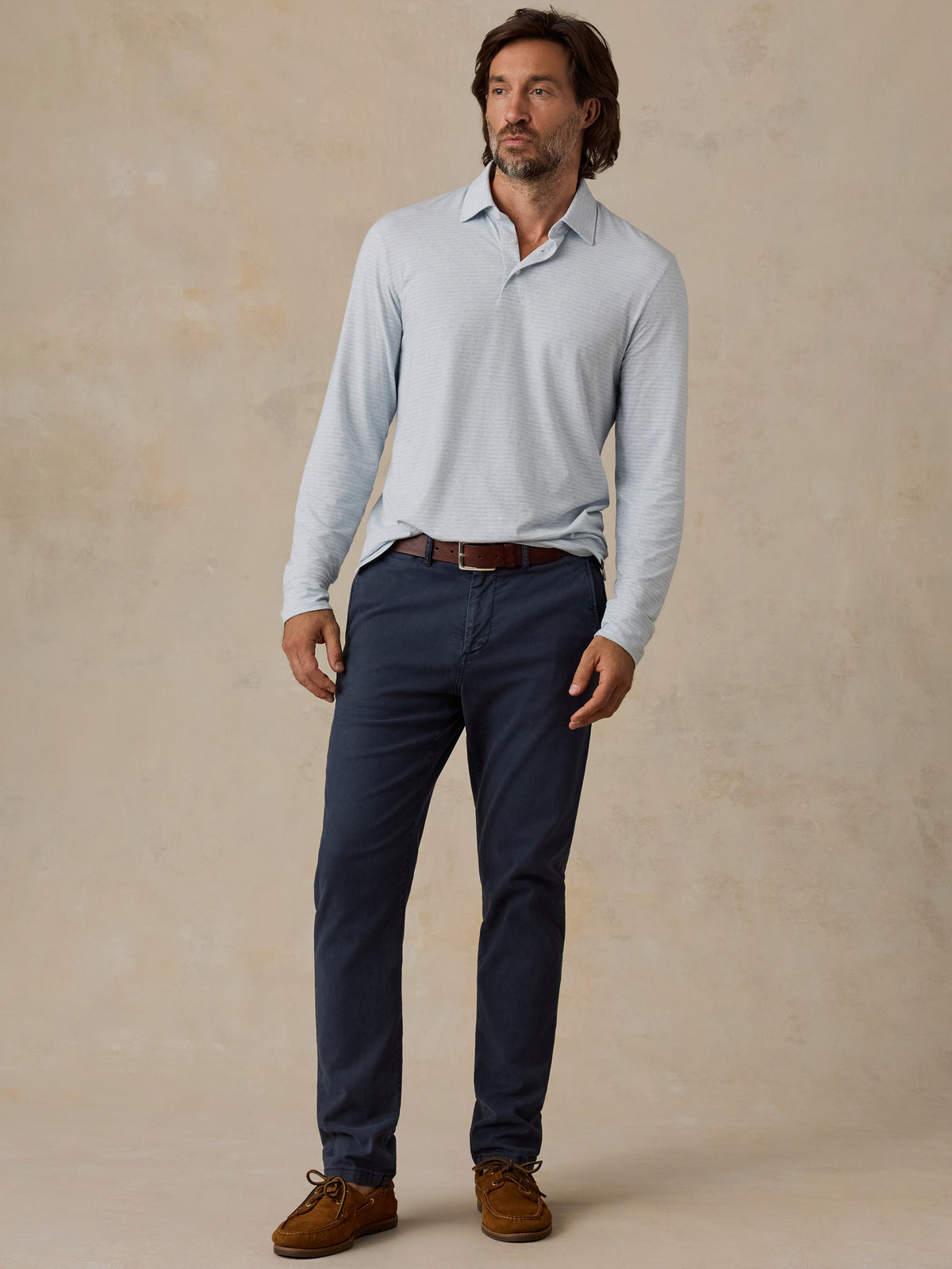 A man modeling a light blue long-sleeve polo shirt, navy blue pants, a brown belt, and brown suede shoes against a textured beige studio backdrop., image 6 of 6