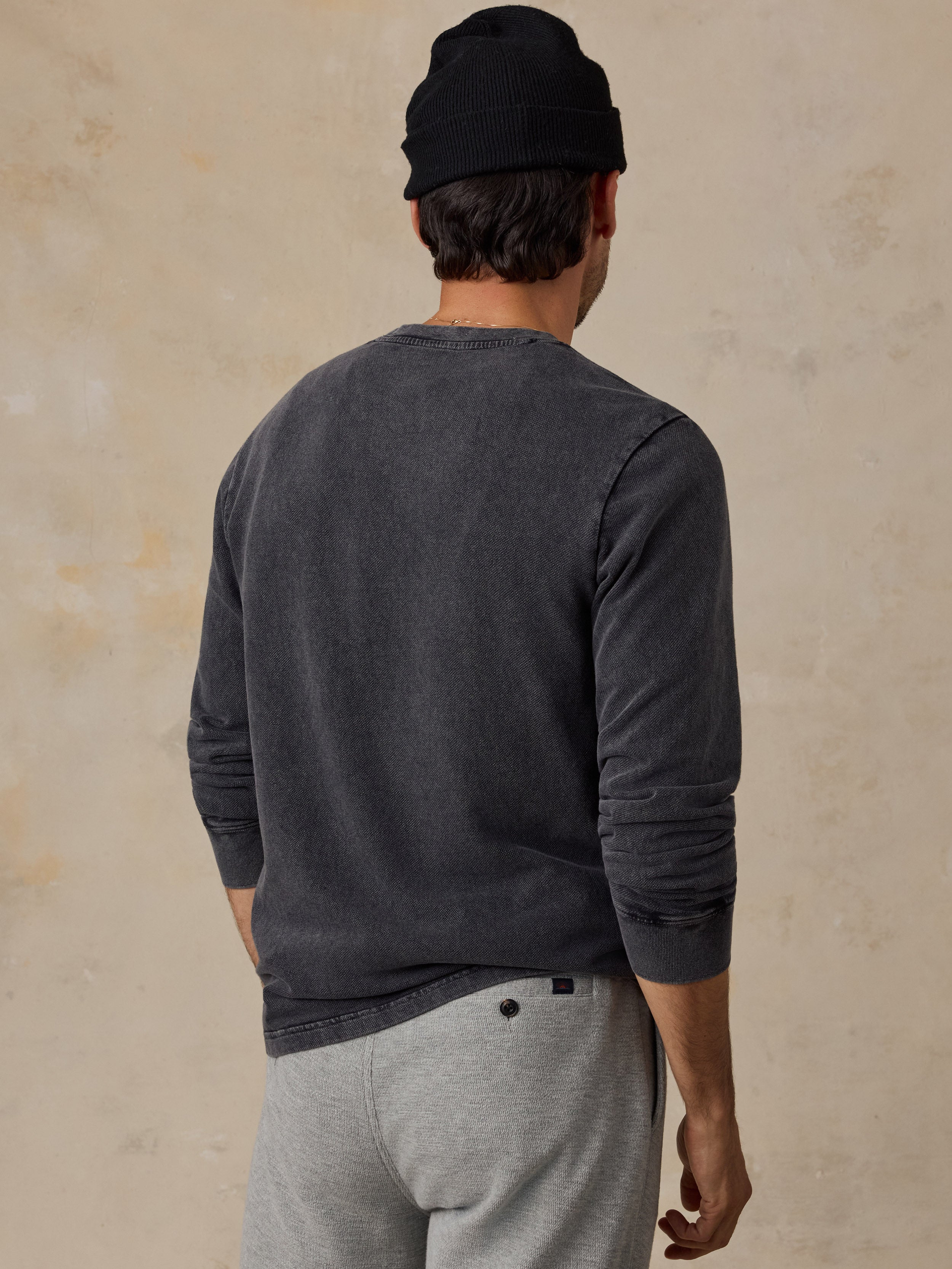 A man viewed from behind wearing a dark charcoal gray long-sleeve crewneck sweater, a black beanie, and light gray sweatpants against a textured beige wall.