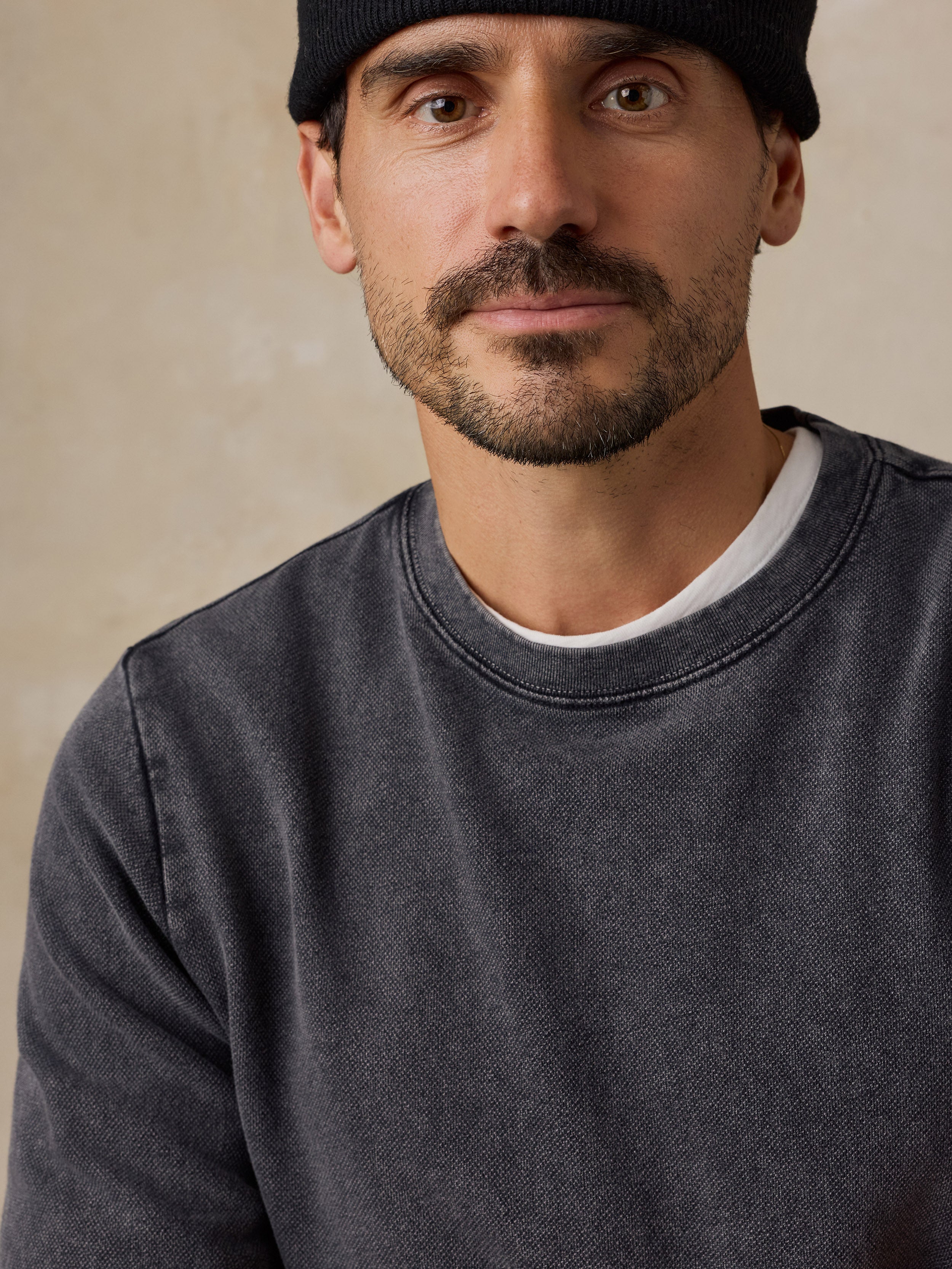 A close-up portrait of a man wearing a dark charcoal gray crewneck sweatshirt over a white undershirt, with a black beanie on a textured beige background.
