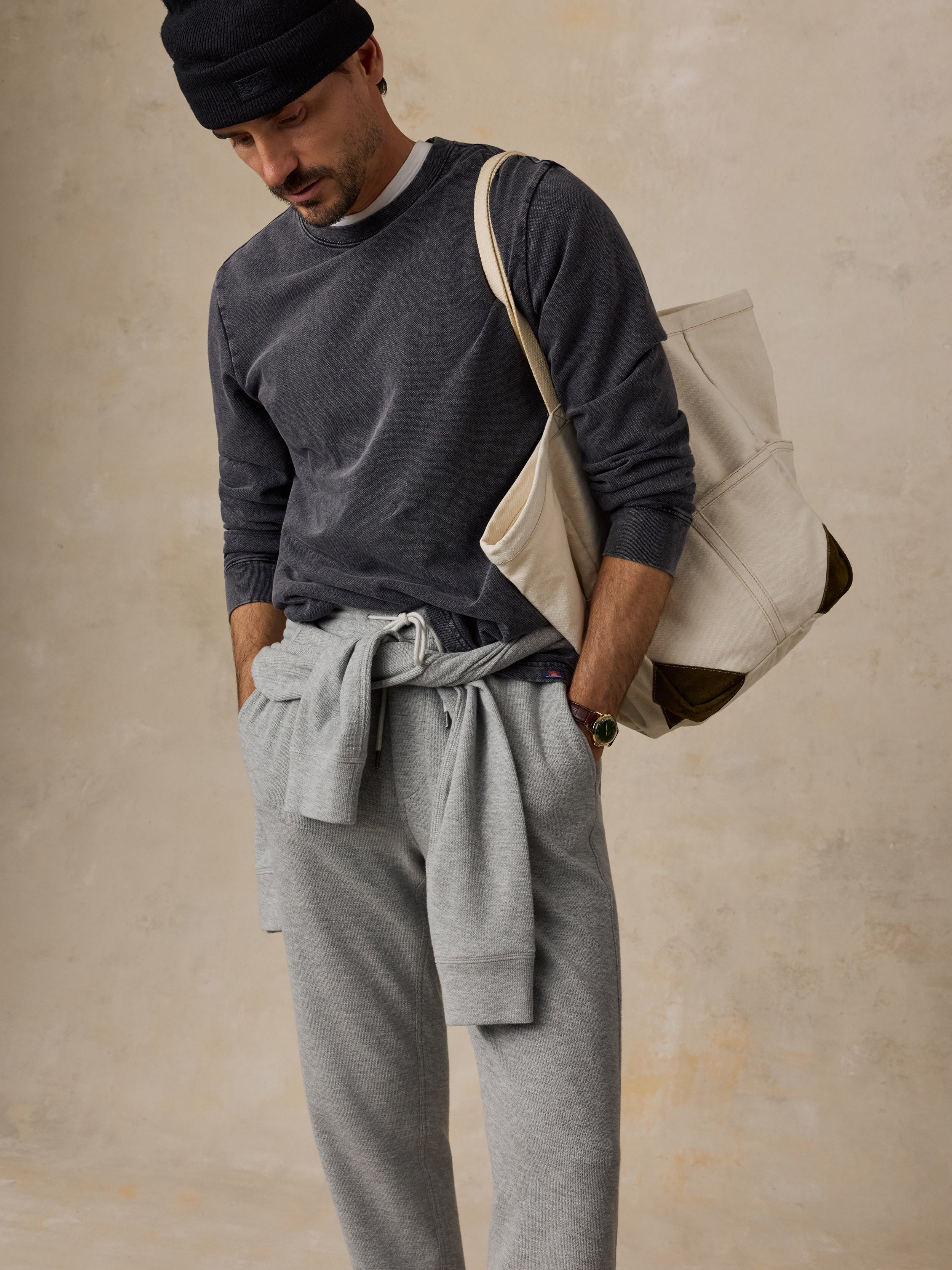 A man wearing a dark gray long-sleeve shirt, light gray sweatpants, a black beanie, and carrying a large cream canvas tote bag over his shoulder, set against a textured beige wall.