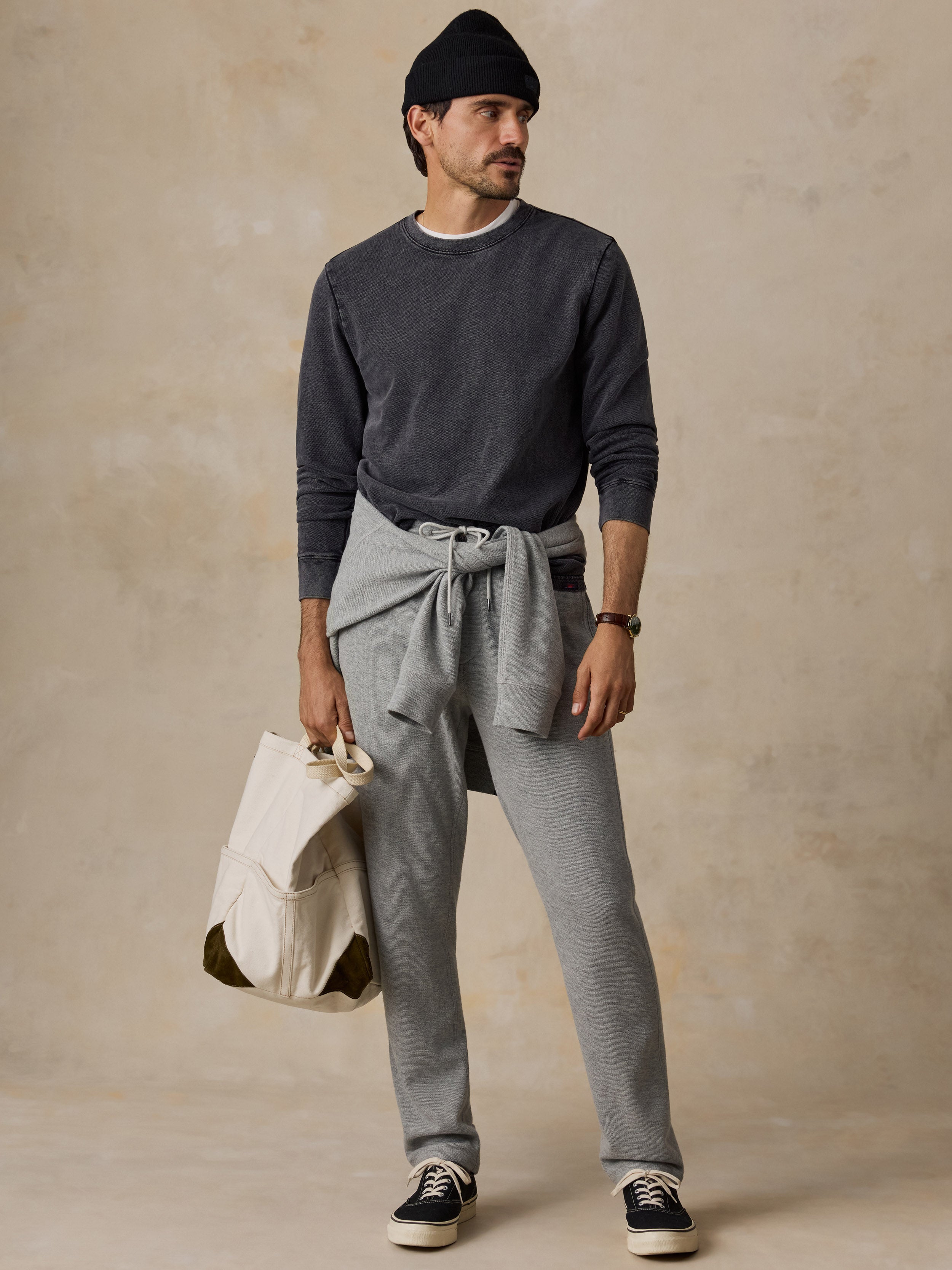 A man modeling a dark charcoal crewneck sweatshirt, light gray tie-front sweatpants, a black beanie, and black sneakers, holding a cream canvas tote bag against a textured beige backdrop.