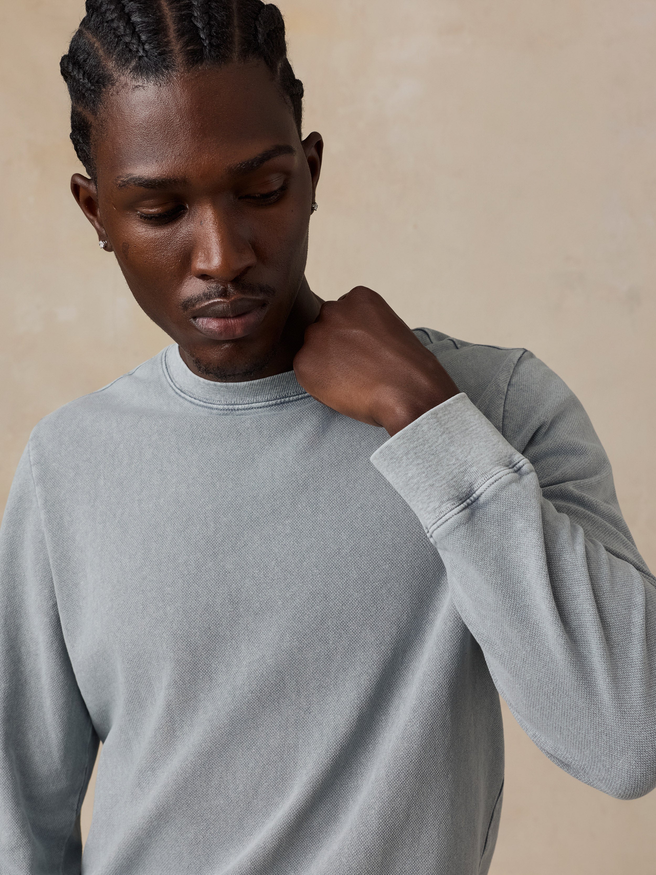A close-up of a man wearing a light gray crewneck sweatshirt, looking down and touching his neck, set against a neutral beige background.