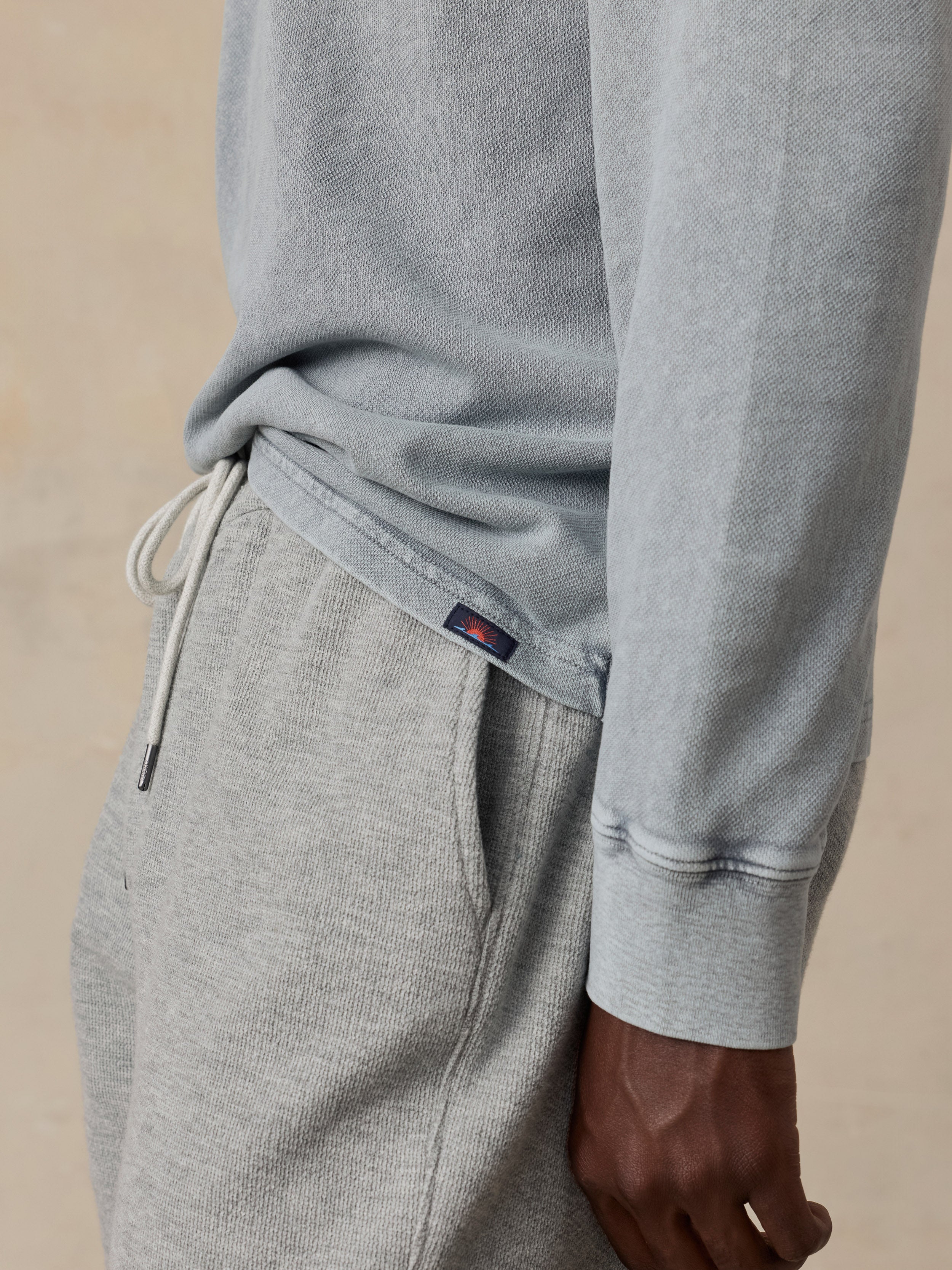 Close-up of a man wearing a light heather gray sweatshirt and matching gray jogger pants, with the drawstring waistband and a small embroidered Faherty brand tab visible.