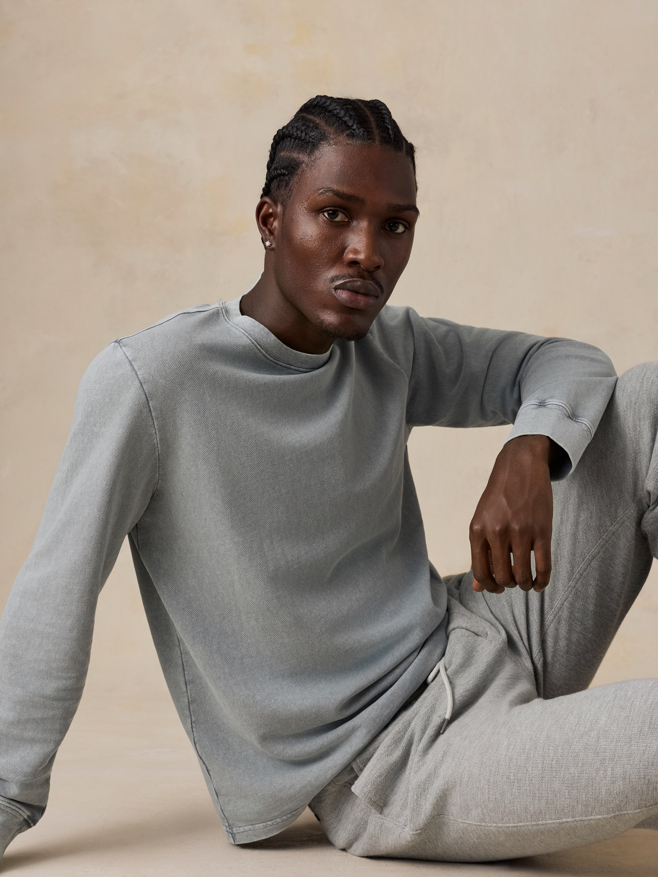A man wearing a light gray long-sleeve crewneck sweatshirt and matching gray sweatpants, sitting against a warm beige textured studio background.