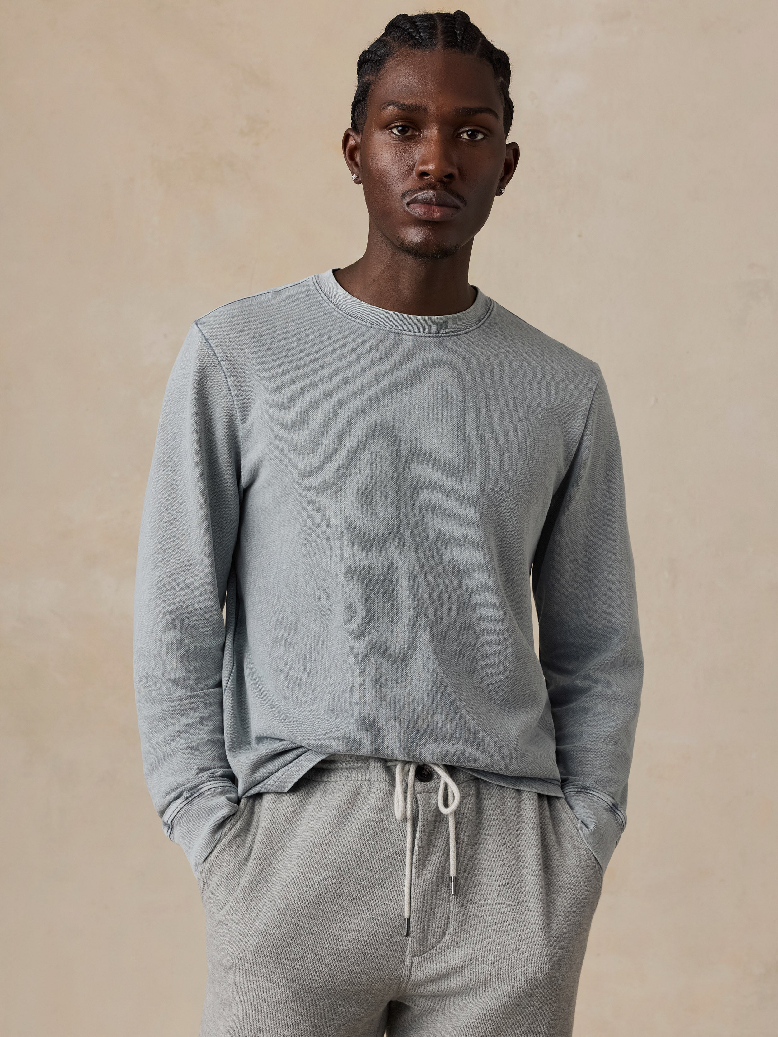 A man wearing a light gray long-sleeve crewneck sweatshirt and matching gray sweatpants, standing against a textured beige background.