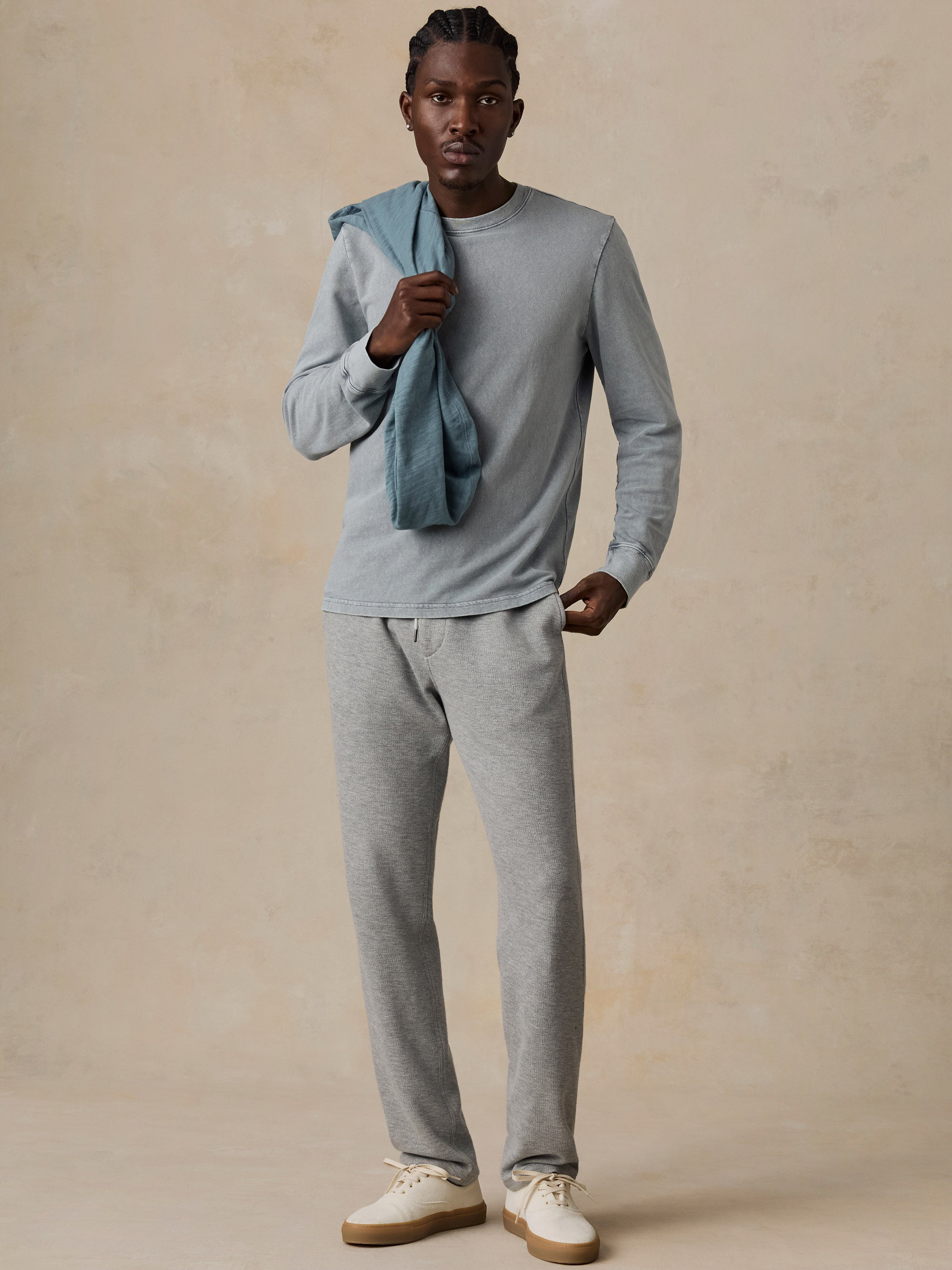 A man modeling a light gray long-sleeve crewneck shirt and matching heather gray sweatpants, with a dusty blue shirt draped over his shoulder, set against a textured beige studio backdrop.
