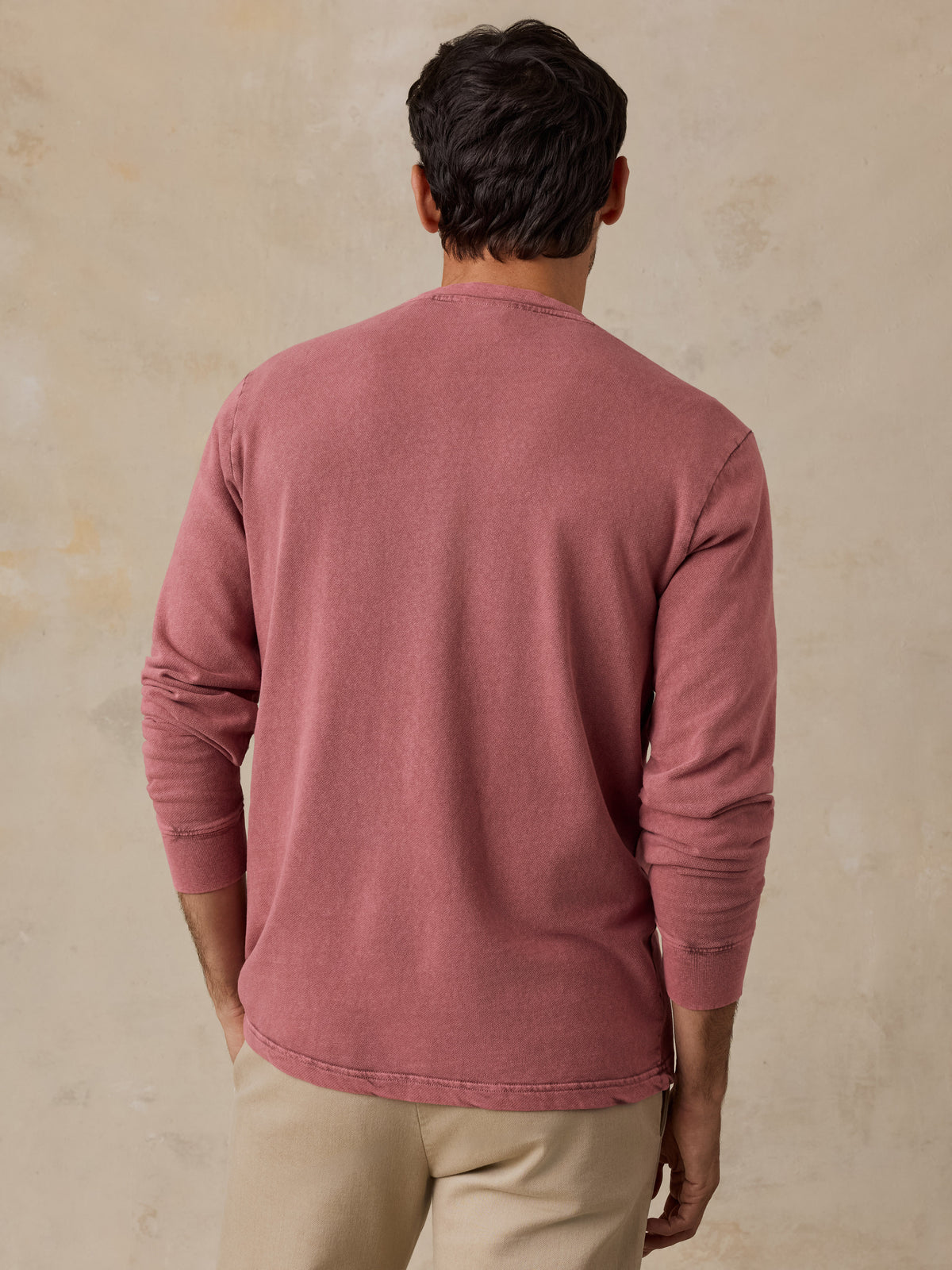 A man viewed from behind wearing a dusty rose long-sleeve shirt and beige pants, standing against a textured beige wall., image 7 of 7